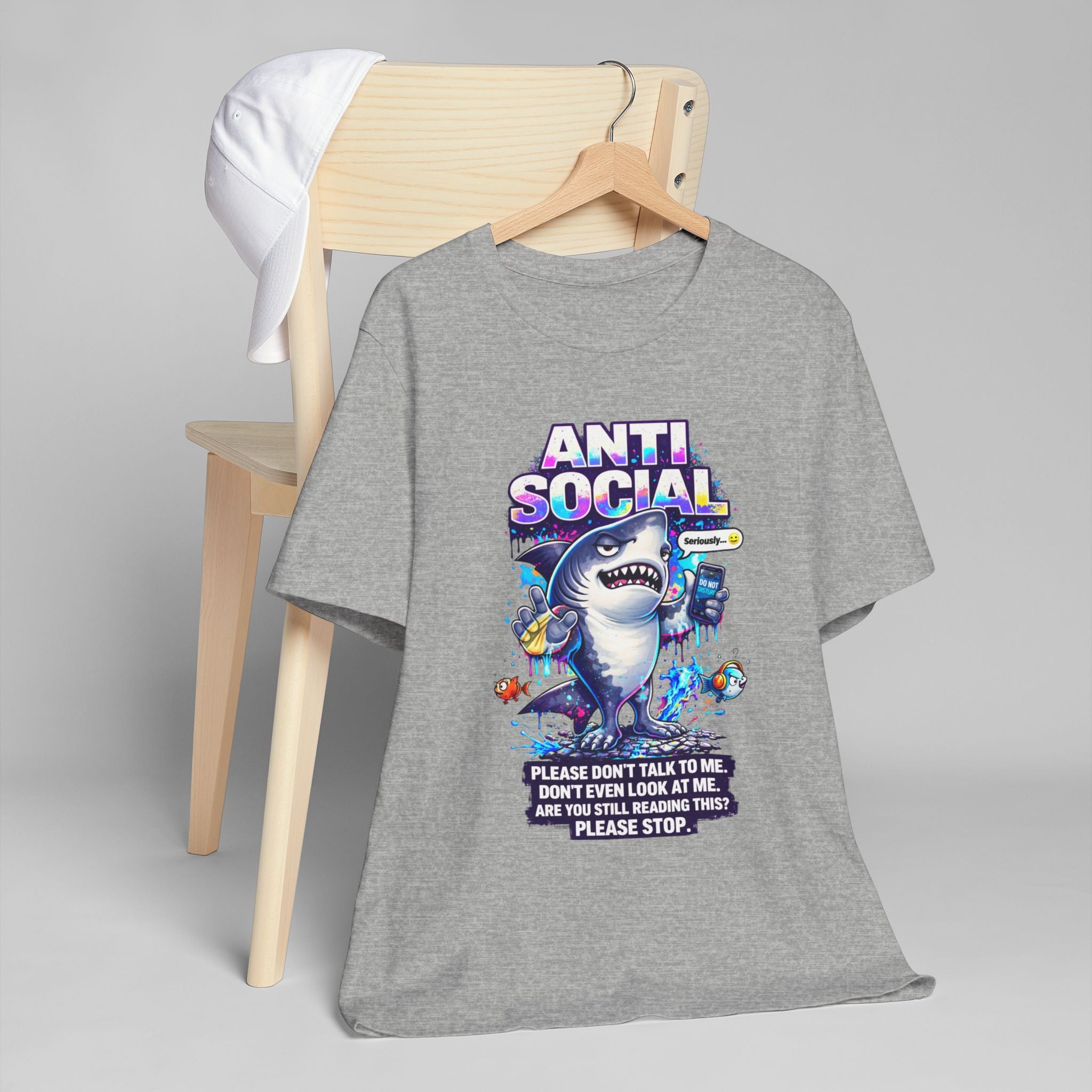 A gray Anti Social Shark T-Shirt — Funny Introvert Graphic Tee with a cartoon shark and colorful graphics hangs on a chair, letting others know to keep their distance. A white cap also rests on the chair.