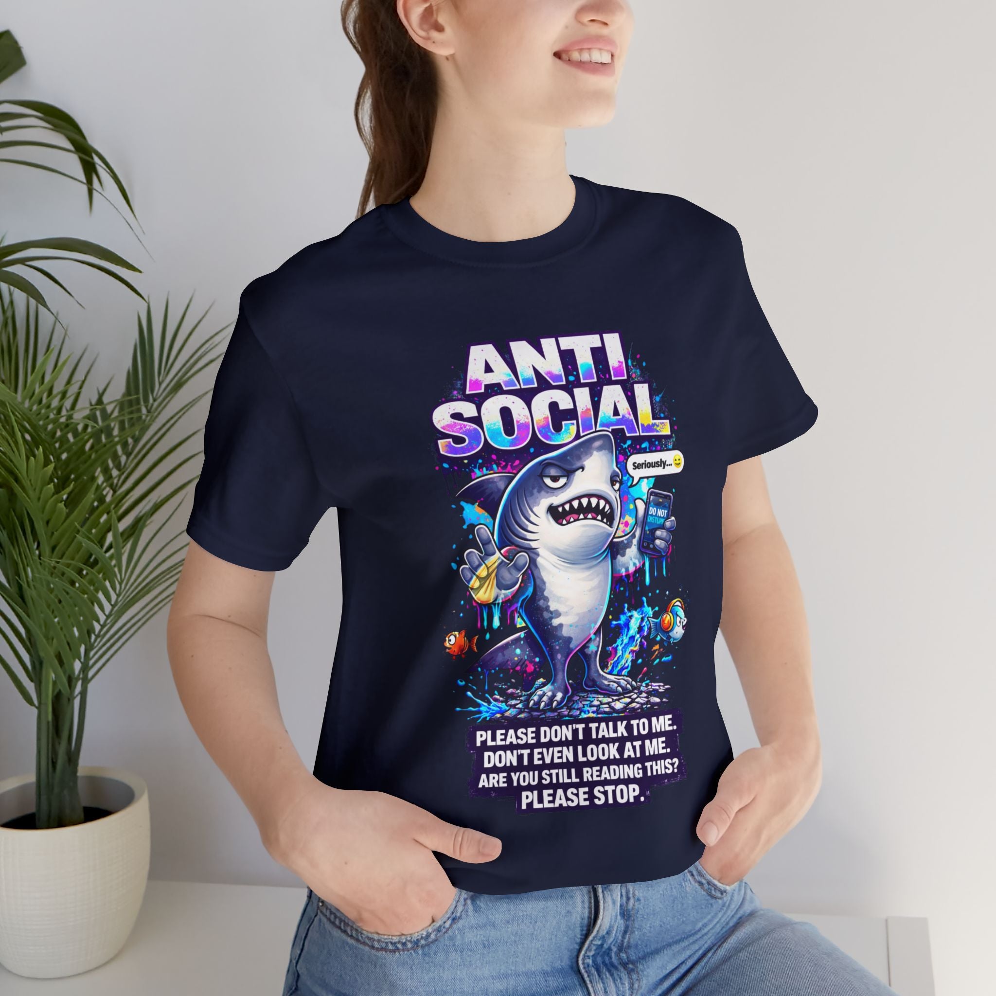 A woman smiles indoors by a plant in the Anti Social Shark T-Shirt — Funny Introvert Graphic Tee, a unisex cotton shirt featuring a cartoon shark with snacks and the slogan, "ANTI SOCIAL. Please don’t talk to me, don’t even look at me.
