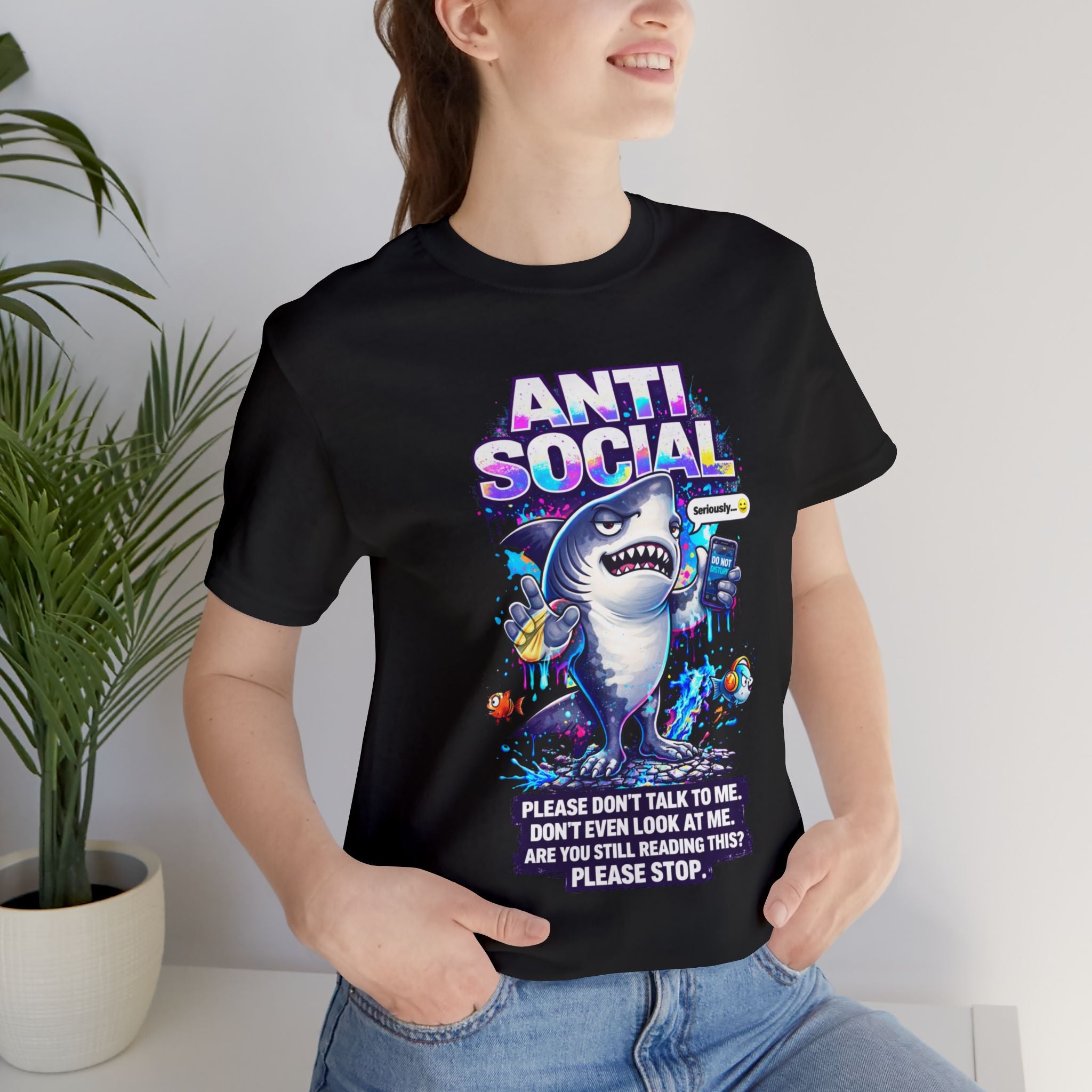 Indoors, a person stands near a green potted plant wearing the Anti Social Shark T-Shirt — Funny Introvert Graphic Tee: a unisex cotton tee with a cartoon shark and colorful "ANTI SOCIAL" text plus funny phrases for introverts.