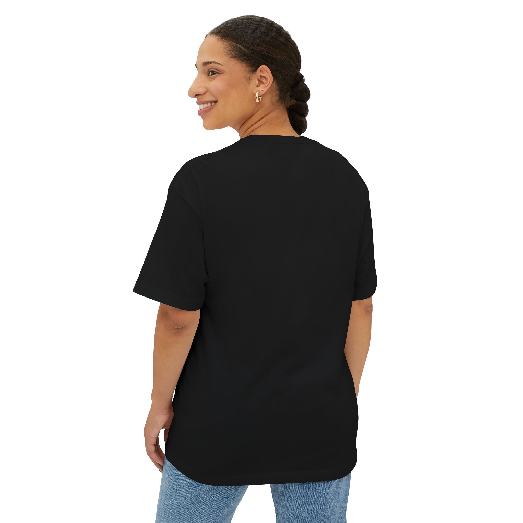 A woman with dark braided hair smiles over her shoulder, wearing the Crowned In Every Era Unisex Oversized Boxy Tee in black (100% Airlume cotton) and blue jeans, standing against a white background.