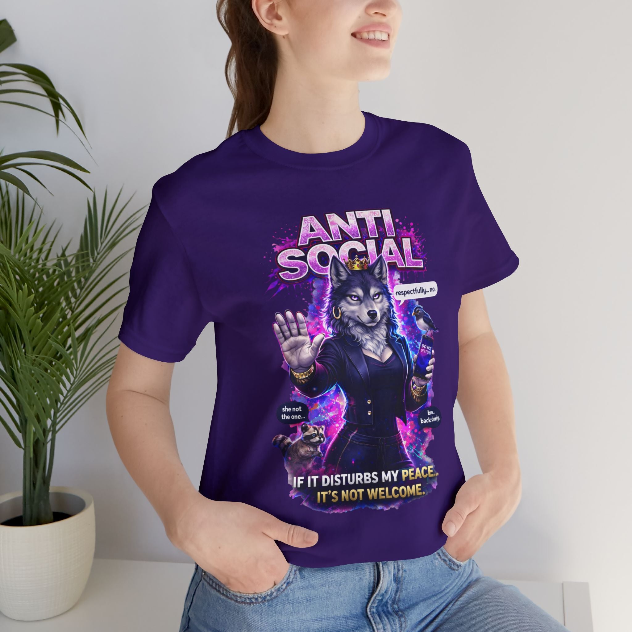 A smiling person stands indoors wearing the Anti Social She-Wolf T-Shirt—Funny Introvert Graphic Tee, featuring a purple design of a wolf in a suit with the phrase: If it disturbs my peace, it’s not welcome. A potted plant sits in the background.