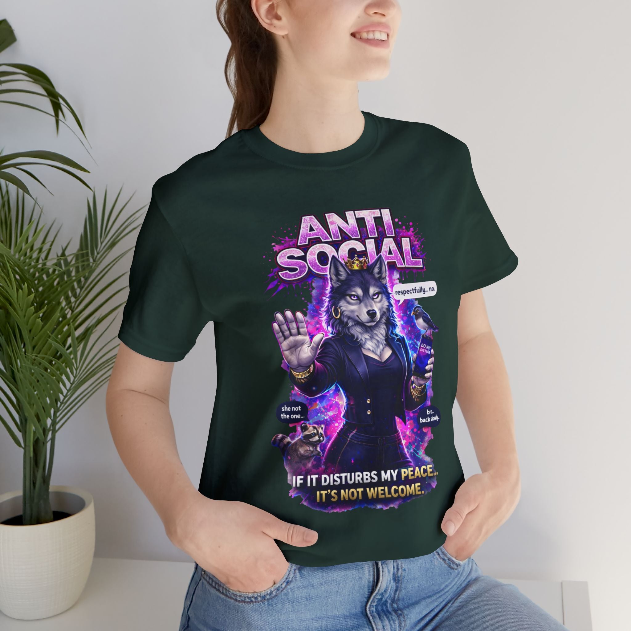 A smiling person wears the Anti Social She-Wolf T-Shirt — a funny introvert graphic tee with a wolf in a suit and sunglasses, bold colors, and the phrase "ANTI-SOCIAL. IF IT DISTURBS MY PEACE, IT'S NOT WELCOME" beside a potted plant.