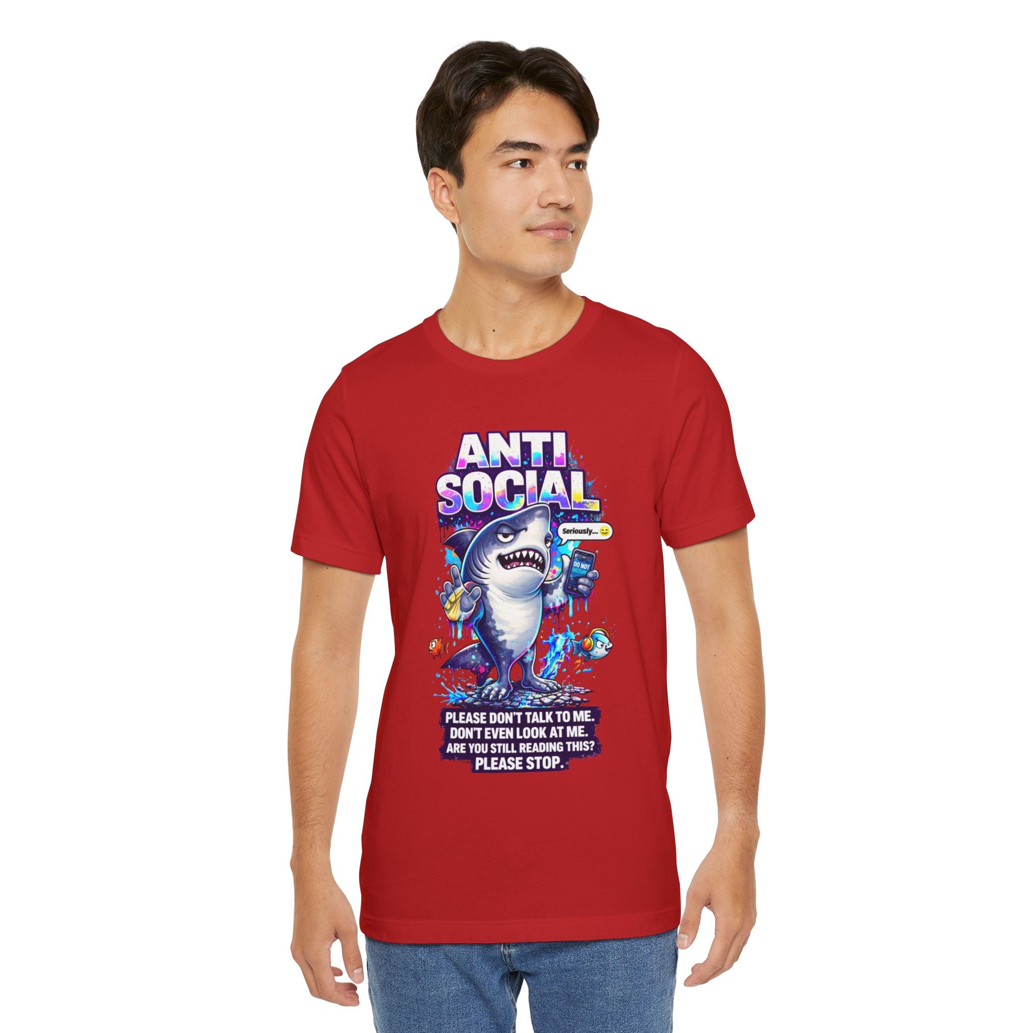 A young man wears the Anti Social Shark T-Shirt — Funny Introvert Graphic Tee, a red unisex cotton shirt with a cartoon shark, fish, bold “ANTI-SOCIAL” text, and the message: Please don’t talk to me. Don’t even look at me. Are you still reading?.