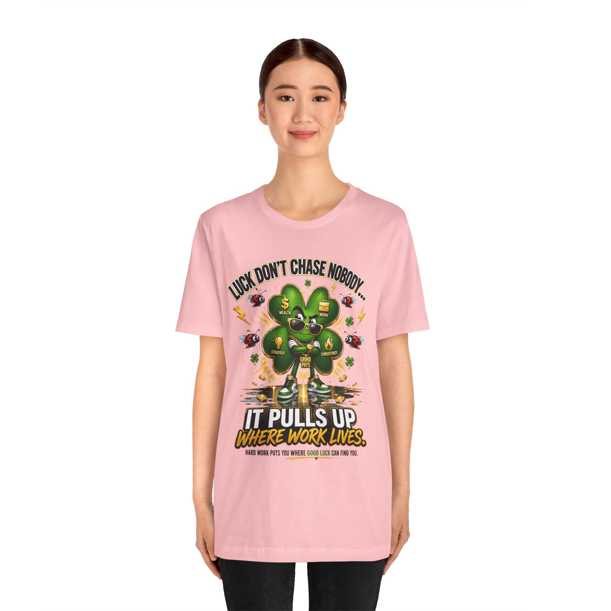 A young woman smiles in a light pink TimeWarp Drip Lucky Clover "Pulls Up Lives" Graphic T-Shirt (S-3XL), featuring a cartoon clover, gold coins, and "Luck don't chase nobody. It pulls up where work lives." Plain white background.