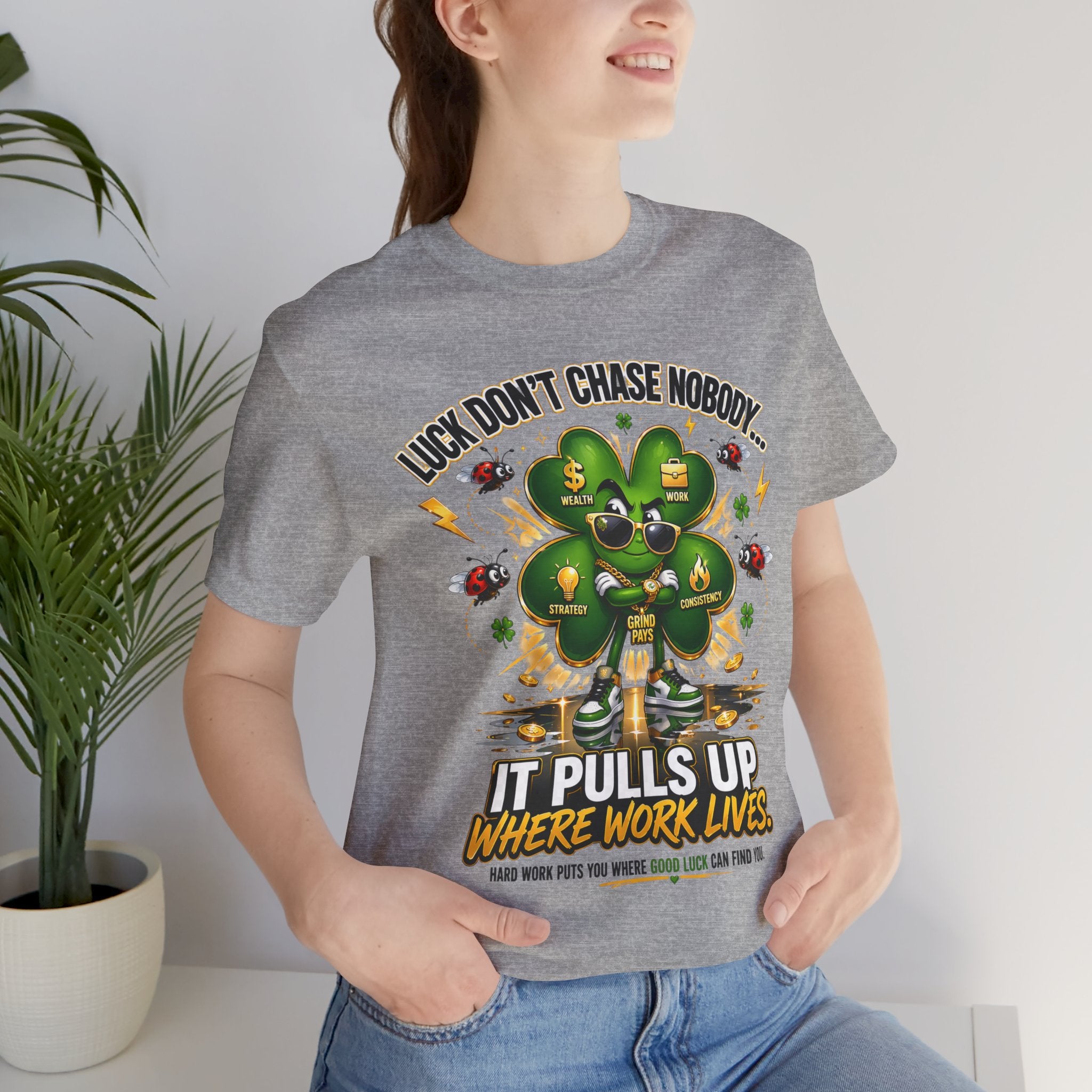 A woman stands indoors, smiling with one hand in her pocket, wearing the TimeWarp Drip Lucky Clover "Pulls Up Lives" Graphic T-Shirt in gray (sizes S-3XL) featuring a clover with cartoon faces and symbols; a potted plant is visible behind her.