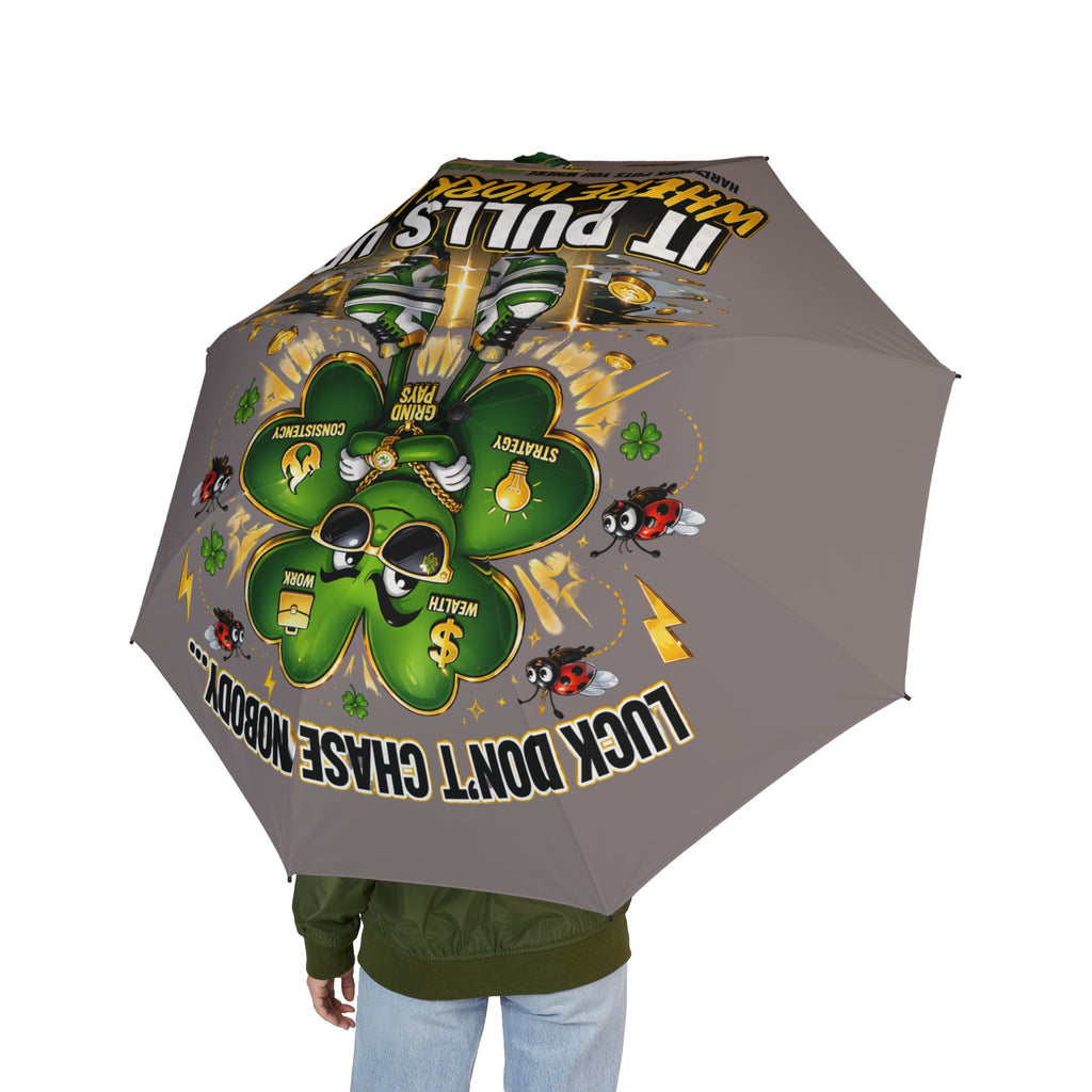 A person holds the Lucky Clover "Pulls Up Lives" grey foldable umbrella featuring a four-leaf clover, cartoon bees, dollar signs, and words like Wealth, Strategy, Consistent Wins. “Look, don’t chase, nobody” is visible on the canopy.