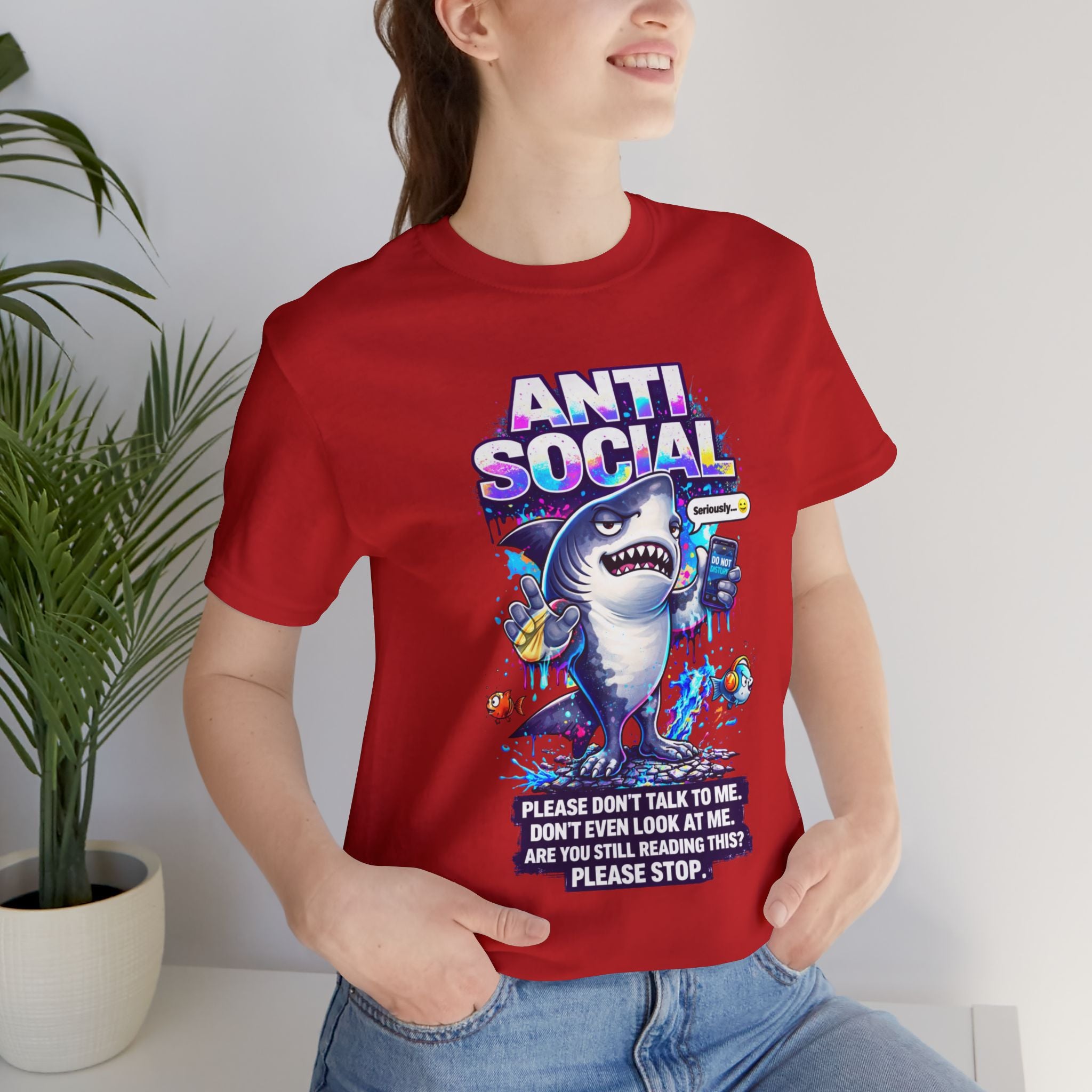 Wearing the Anti Social Shark T-Shirt — Funny Introvert Graphic Tee, a person smiles next to a potted plant. The unisex cotton shirt features a cartoon shark and playful text: Please don’t talk to me or even look at me.