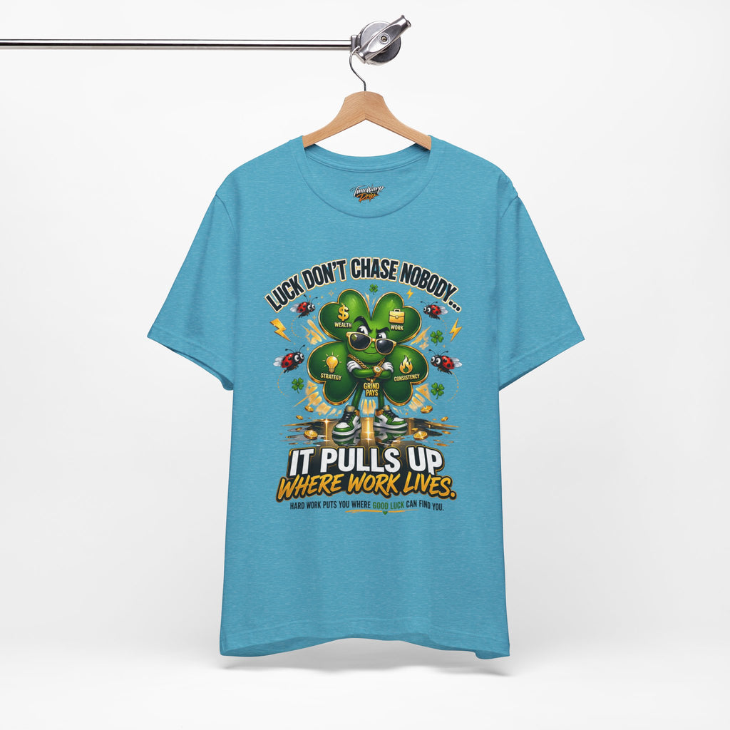 The Lucky Clover "Pulls Up Lives" Graphic T-Shirt by TimeWarp Drip in light blue (sizes S-3XL) hangs on a wooden hanger, featuring a four-leaf clover with money symbols and the phrase: Luck don’t chase nobody. It pulls up where work lives.