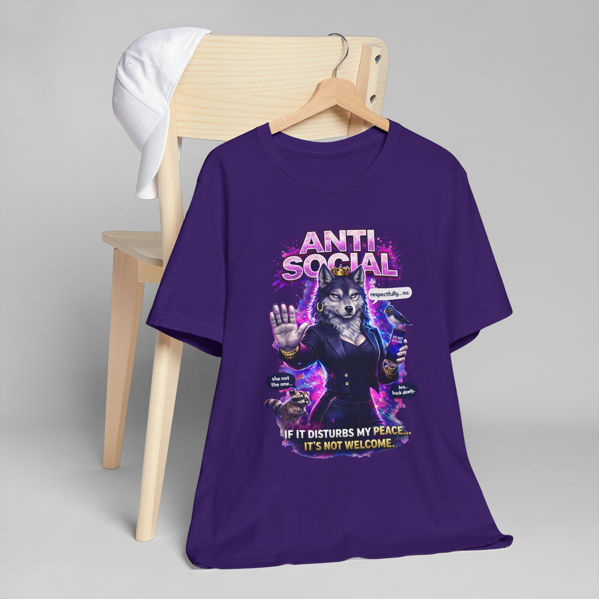 The Anti Social She-Wolf T-Shirt — a purple introvert tee with a suited wolf graphic and the text “ANTI SOCIAL” and “If it disturbs my peace... it’s not welcome.” Displayed draped over a wooden chair with a white cap.