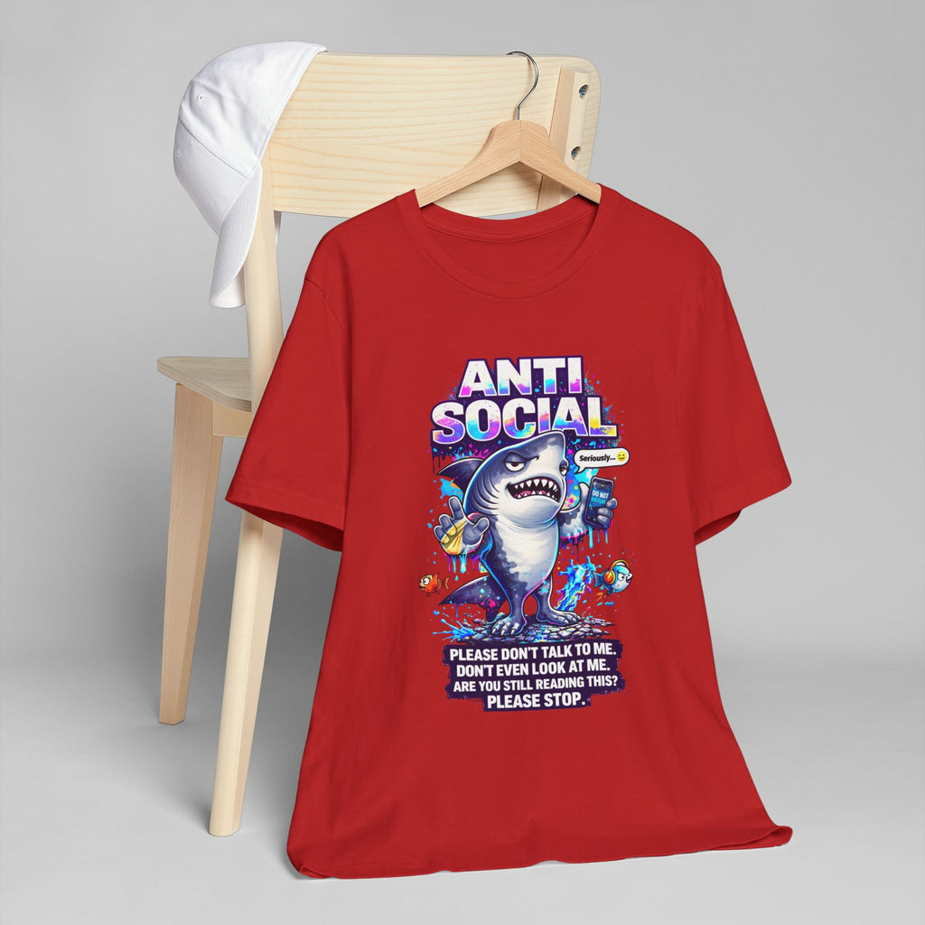 The Anti Social Shark T-Shirt — Funny Introvert Graphic Tee shows a red shirt with a cartoon shark holding a phone, snacks, and drink amid sea creatures. The unisex cotton tee features "ANTI SOCIAL" text and is displayed on a wooden chair with a white cap.