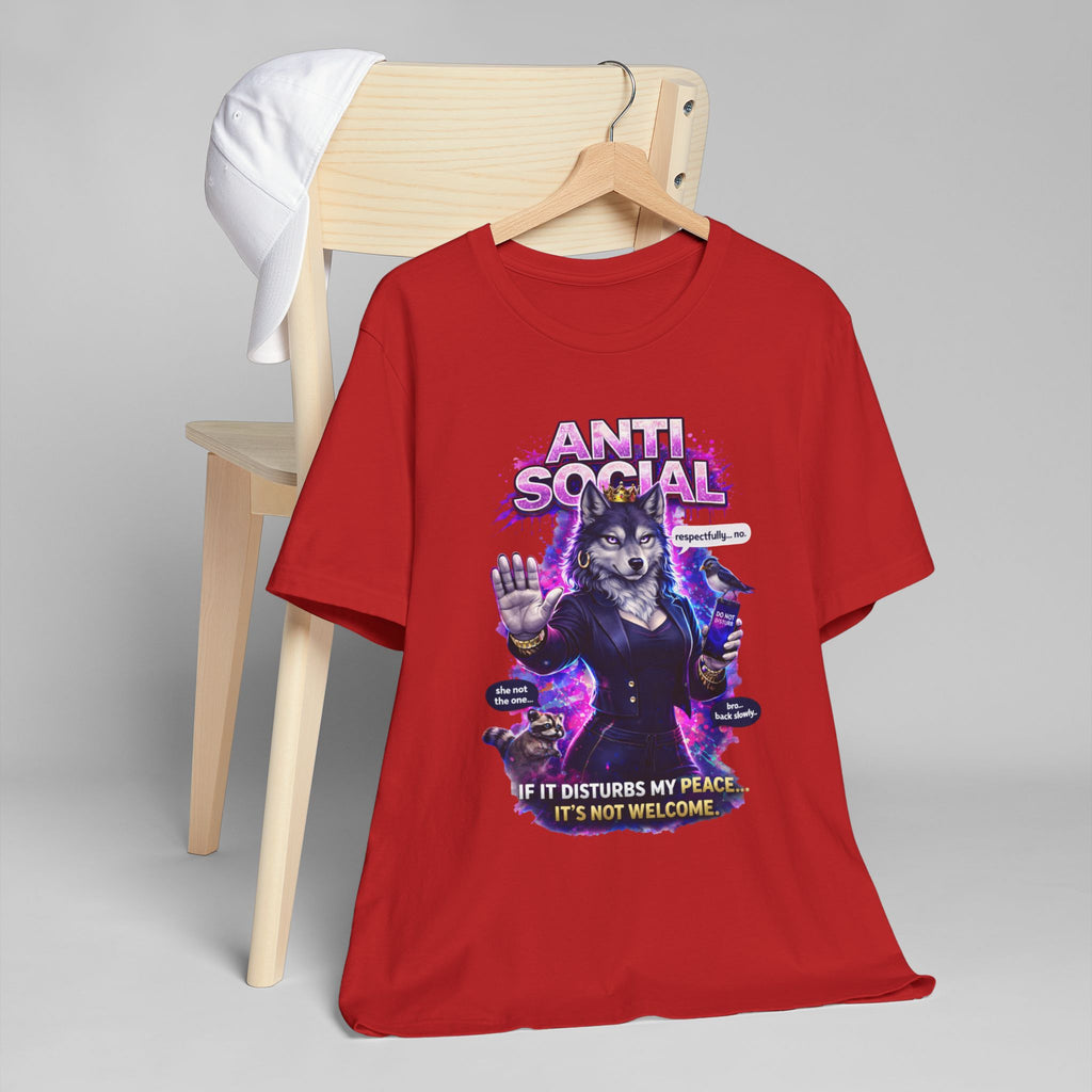 The Anti Social She-Wolf T-Shirt — Funny Introvert Graphic Tee features a grumpy wolf in a hoodie with a phone and the phrases "ANTI SOCIAL" and "If it disturbs my peace… it's not welcome." Pictured on a chair with a white cap.