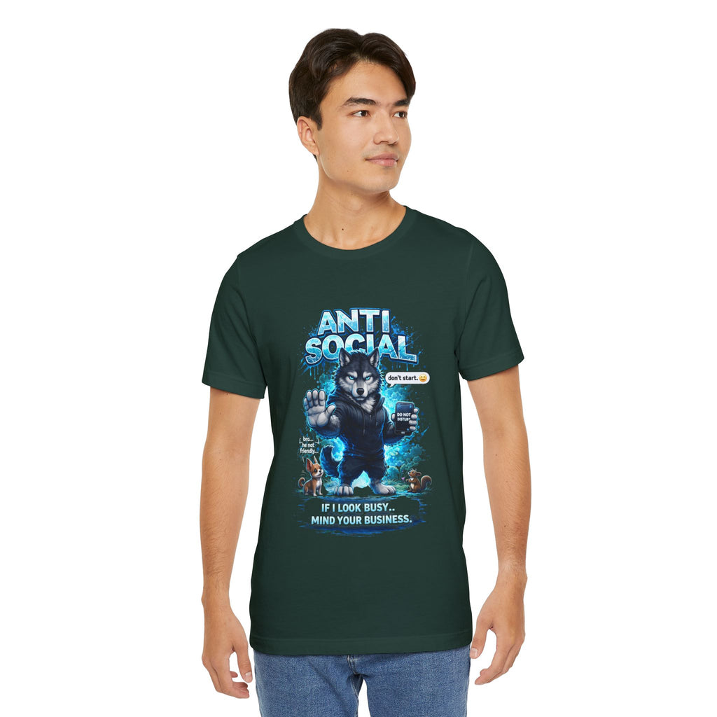 A young man in a dark green Anti Social Wolf T-Shirt — Funny Introvert Graphic Tee, with a grumpy wolf, blue "ANTI SOCIAL" text, and "IF I LOOK BUSY, MIND YOUR BUSINESS," stands against a plain background looking to his left.