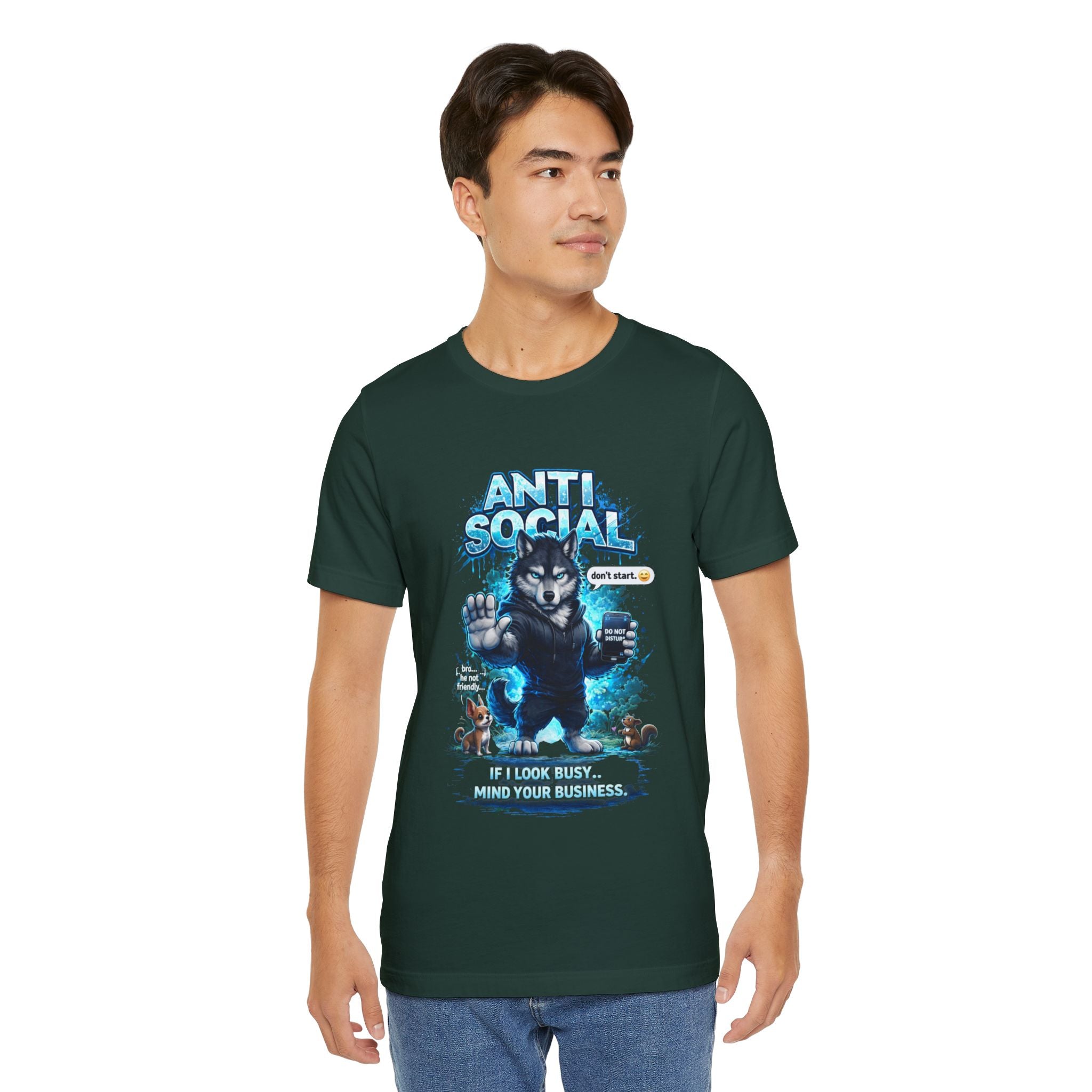 A young man in a dark green Anti Social Wolf T-Shirt — Funny Introvert Graphic Tee, with a grumpy wolf, blue "ANTI SOCIAL" text, and "IF I LOOK BUSY, MIND YOUR BUSINESS," stands against a plain background looking to his left.