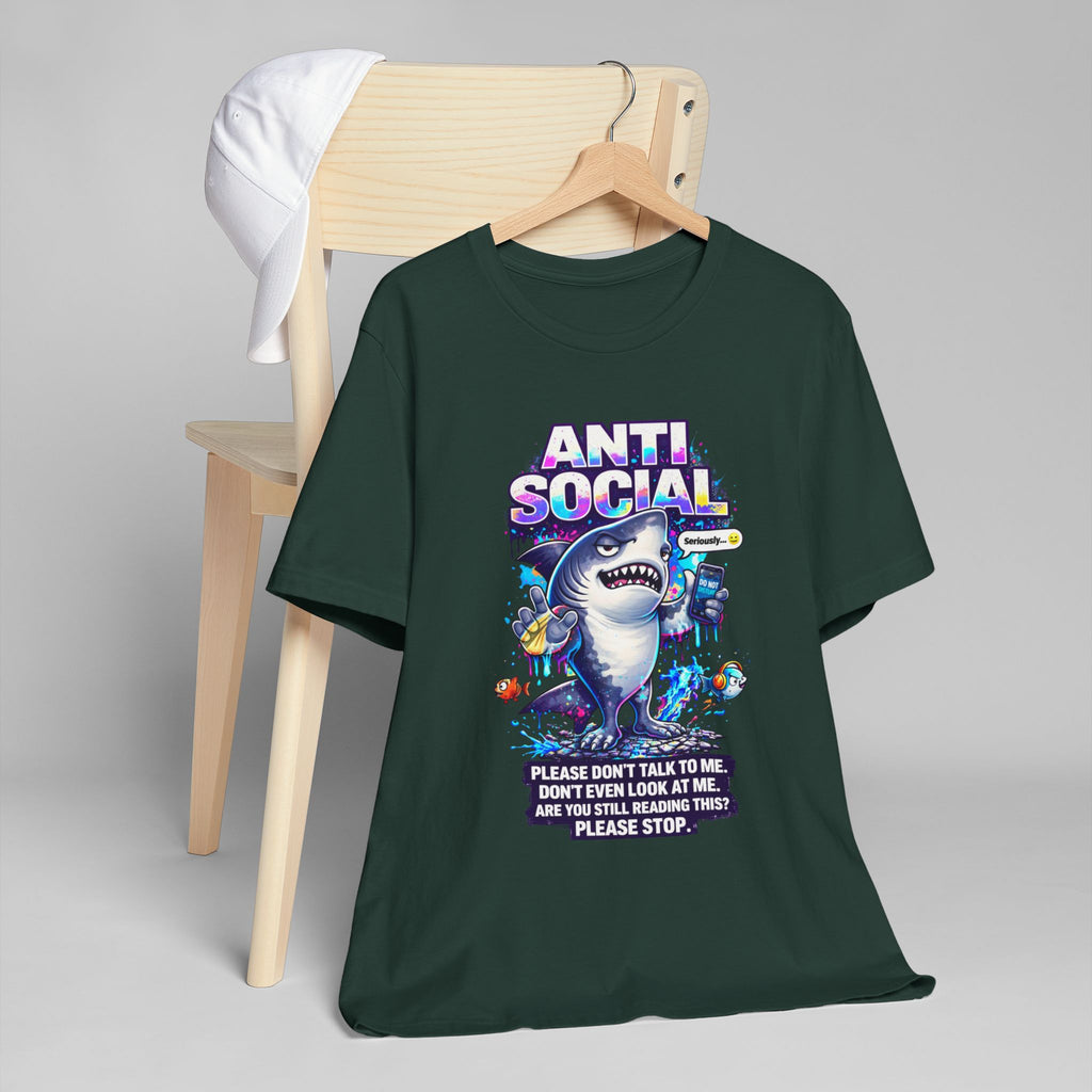 The Anti Social Shark T-Shirt — Funny Introvert Graphic Tee is a dark green unisex cotton tee with a playful shark graphic and colorful accents, displayed on a wooden chair with a white cap. It reads: "Please don't talk to me. Don't even look at me.