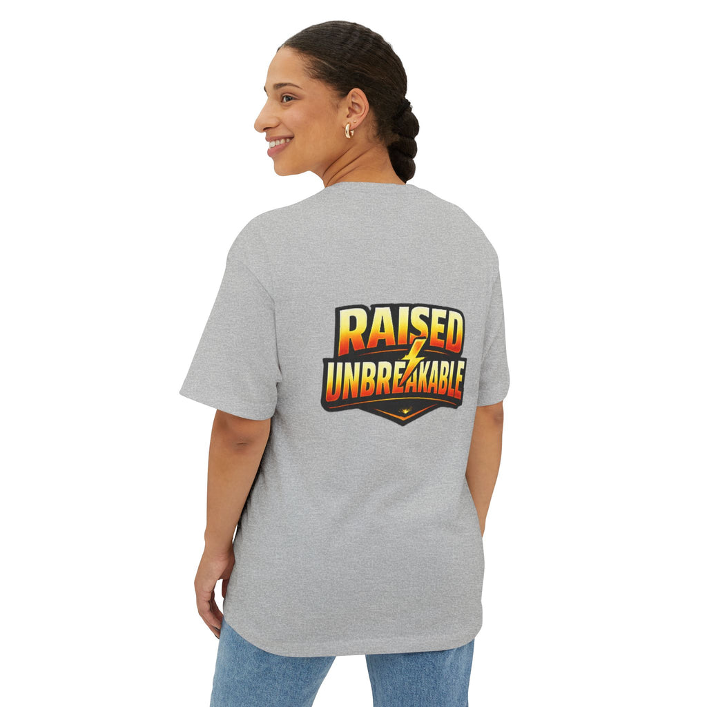 A woman with braided hair smiles over her shoulder, wearing the TimeWarp Drip Generation X Parenting "Come Get Em" oversized boxy tee in gray cotton, featuring bold “RAISED UNBREAKABLE” text on the back in black, yellow, and red. She pairs it with blue jeans.