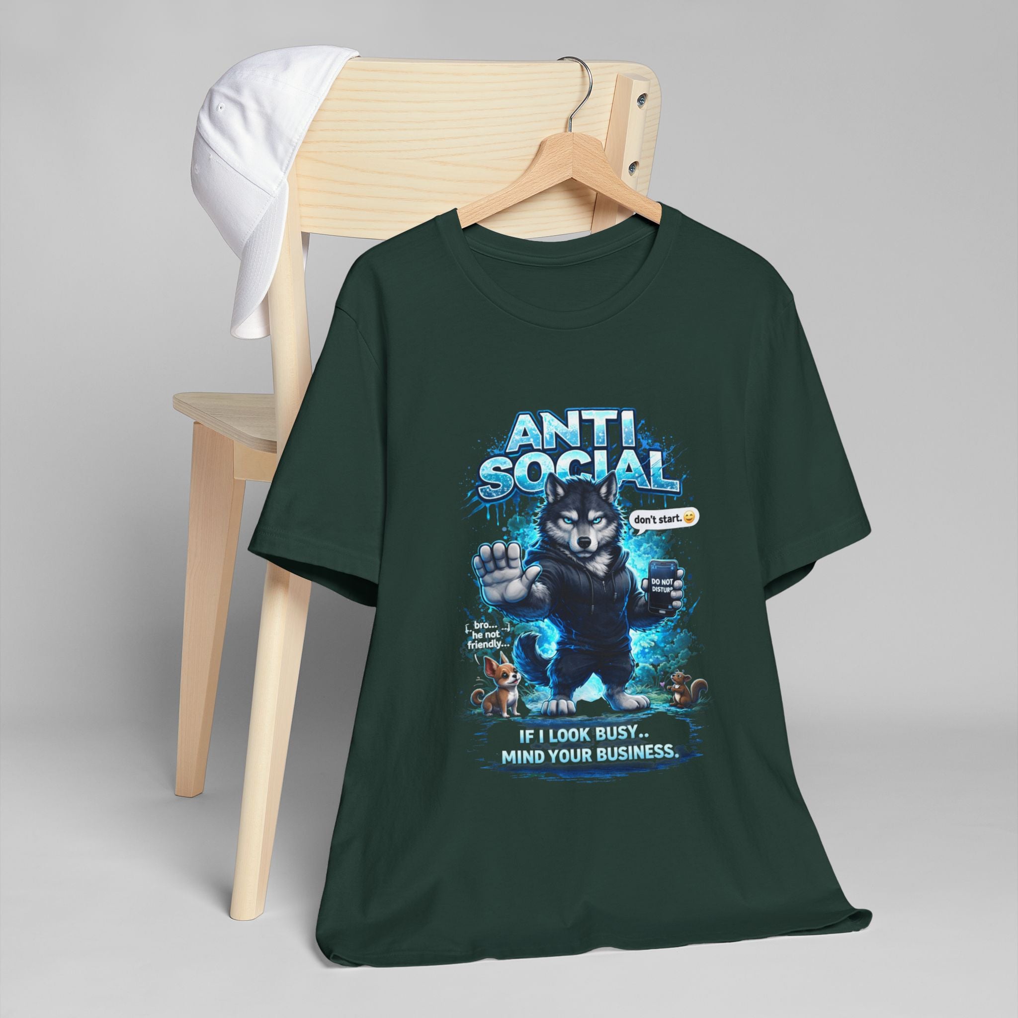 The Anti Social Wolf T-Shirt — Funny Introvert Graphic Tee in dark green features a tough wolf graphic and bold “ANTI SOCIAL” with “If I look busy… mind your business,” draped over a wooden chair topped with a white cap.