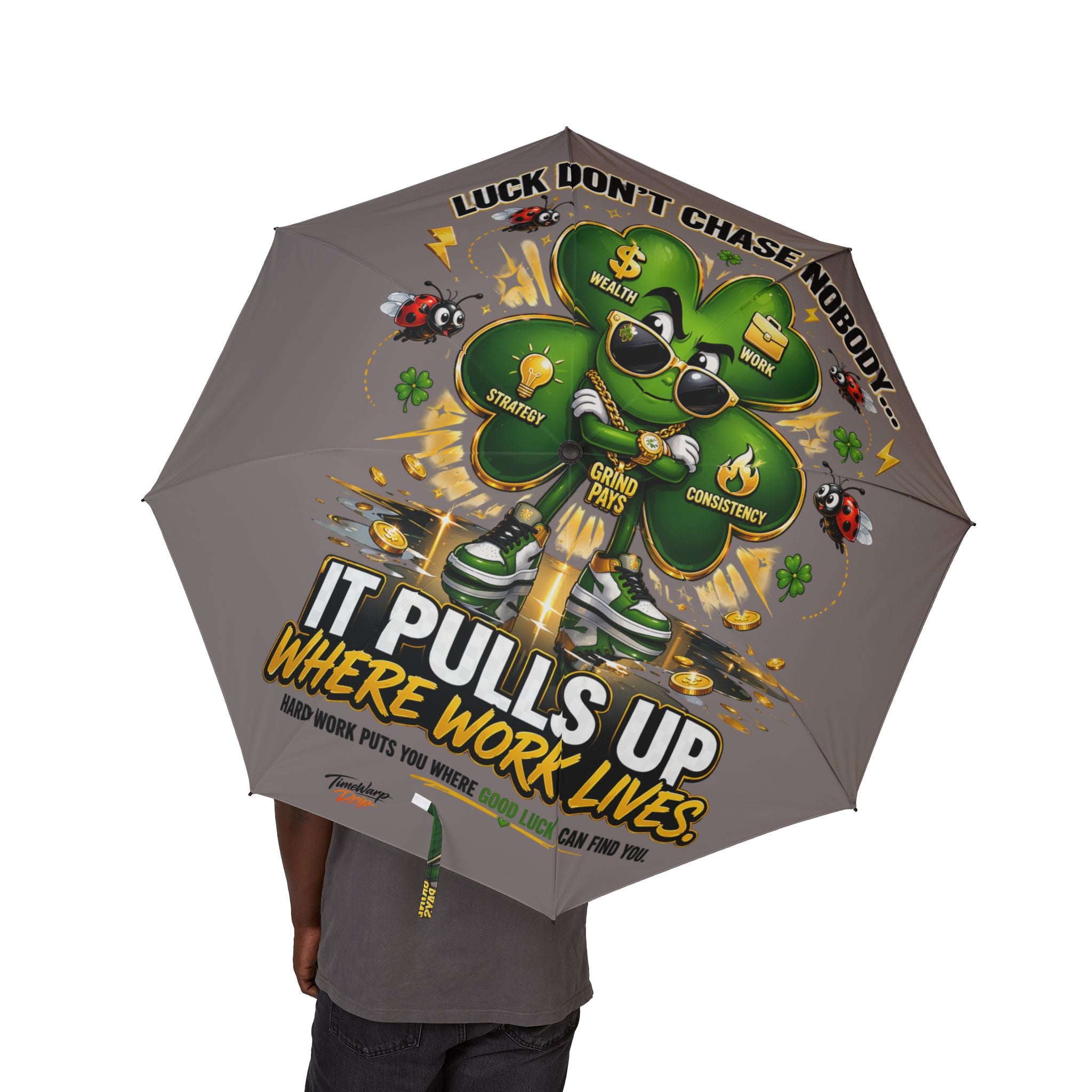 A person holds the Lucky Clover "Pulls Up Lives" Graphic (Grey) Foldable Umbrella, featuring a cartoon shamrock in sneakers with coins and lightning. Motivational graphics pop on gray; one size fits all with an optimal canopy diameter for any occasion.