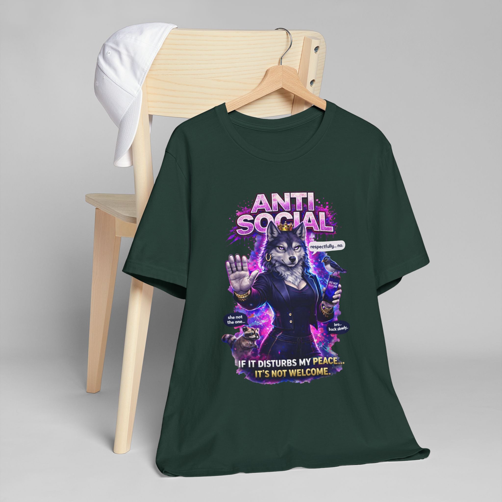 A dark green Anti Social She-Wolf T-Shirt — Funny Introvert Graphic Tee hangs on a wooden chair with a white cap. The tee shows a wolf in a suit, “ANTI SOCIAL” text, and the phrase “If it disturbs my peace... it’s not welcome.”.