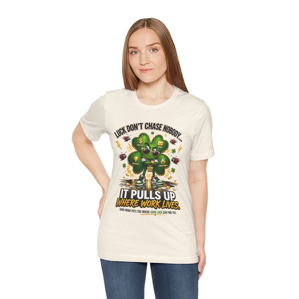 A woman with straight light brown hair wears Lucky Clover "Pulls Up Lives" Graphic T-Shirt, featuring a four-leaf clover and gold coins. Available in sizes S–3XL, the shirt reads: “Luck don’t chase nobody. It pulls up where work lives.”.
