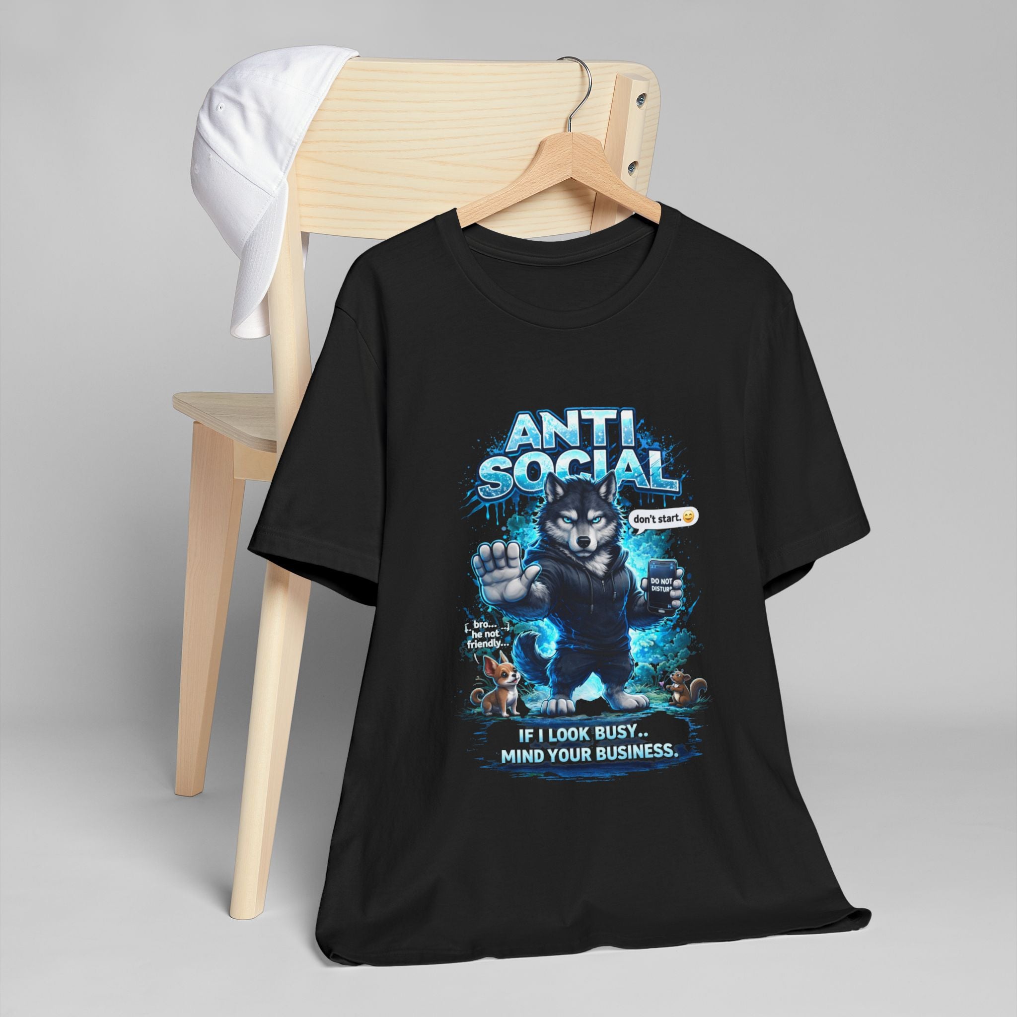 The Anti Social Wolf T-Shirt — Funny Introvert Graphic Tee in black, with a grumpy wolf and “If I look busy… mind your business” text, is draped over a wooden chair topped with a white baseball cap. A must-have for streetwear fans.
