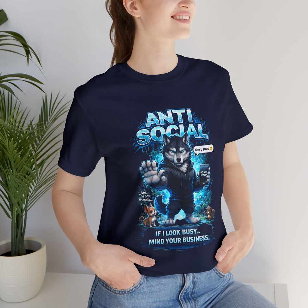A smiling woman stands indoors near a plant, wearing Timewarp Drip’s Anti Social Wolf T-Shirt — Funny Introvert Graphic Tee featuring a cartoon wolf, two small dogs, a "Dont stare" sign, and reads: ANTI SOCIAL and If I look busy... Mind your business.