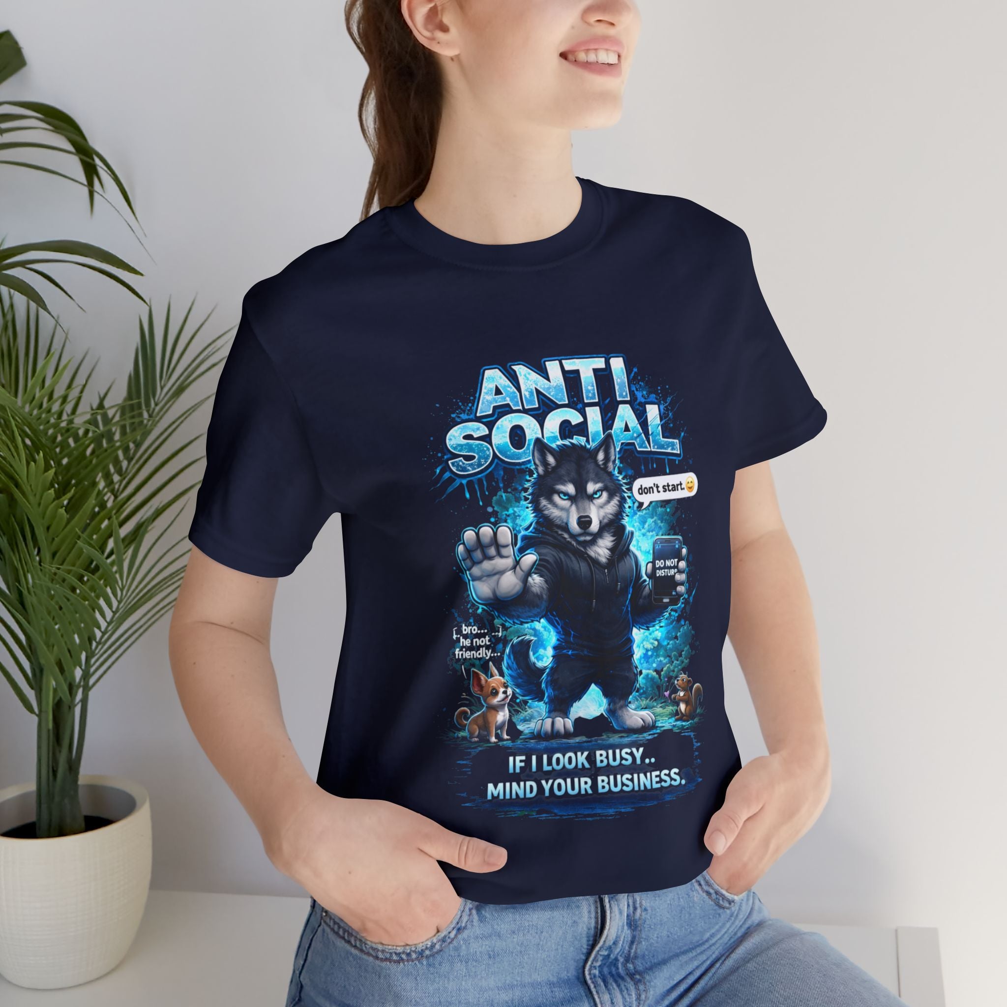A smiling woman stands indoors near a plant, wearing Timewarp Drip’s Anti Social Wolf T-Shirt — Funny Introvert Graphic Tee featuring a cartoon wolf, two small dogs, a "Dont stare" sign, and reads: ANTI SOCIAL and If I look busy... Mind your business.