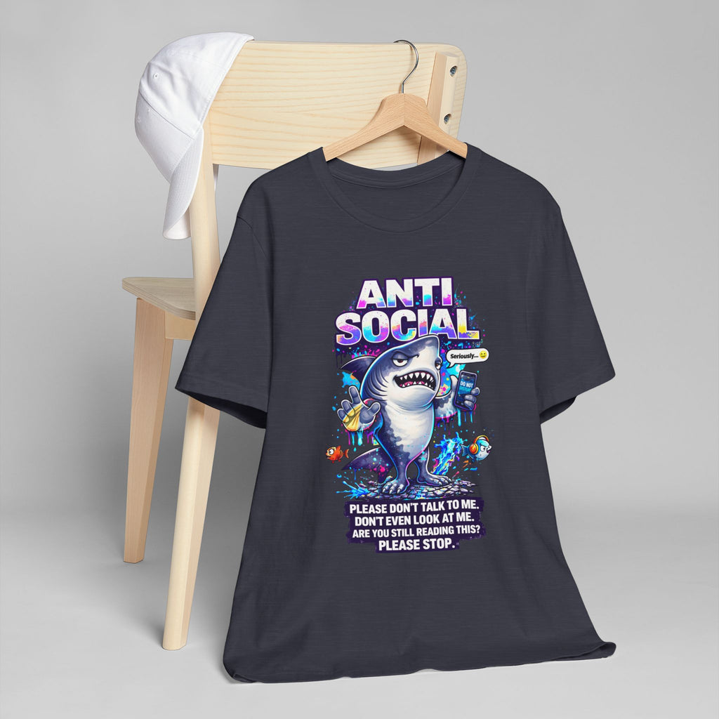 A black unisex cotton Anti Social Shark T-Shirt — Funny Introvert Graphic Tee, featuring a cartoon shark and ‘ANTI SOCIAL’ text with “Please don't talk to me. Don't even look at me.” hangs on a wooden chair. A white cap rests nearby.