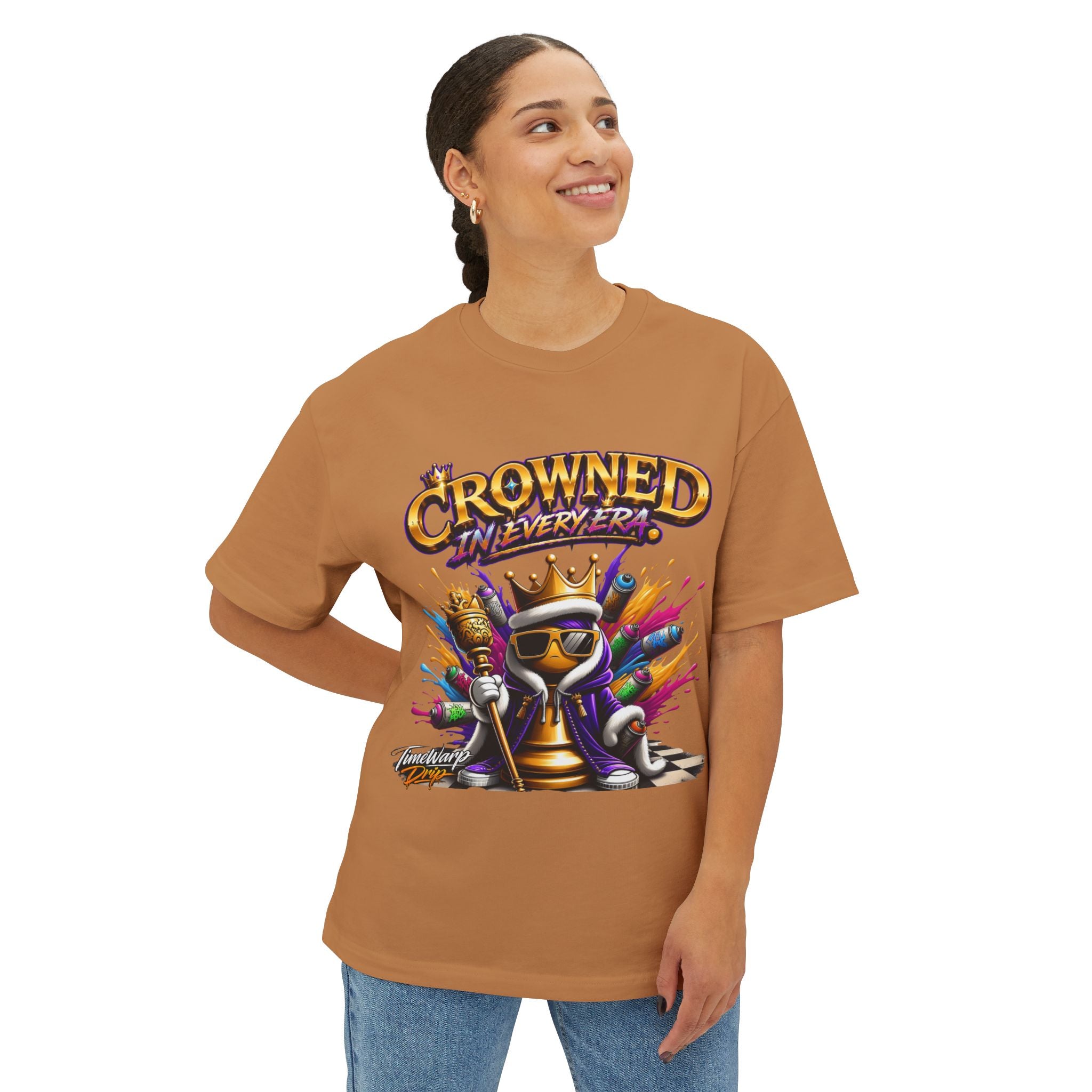 A smiling person wears the Crowned In Every Era - Unisex Oversized Boxy Tee, a tan relaxed fit shirt with a cartoon frog wearing a crown, sunglasses, and scepter amid colorful paint splashes. "CROWNED IN EVERY ERA" is printed in bold letters.