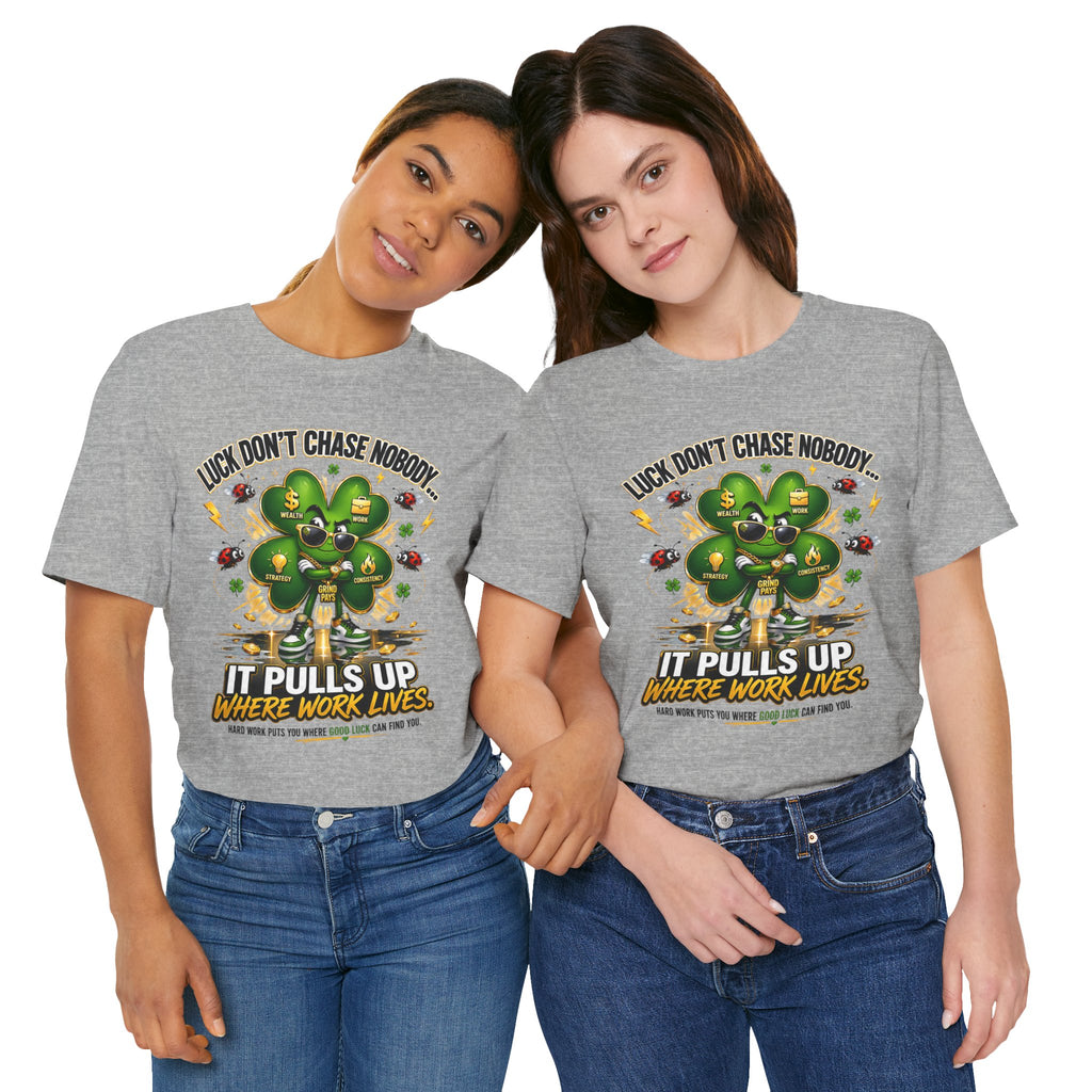 Two women smile and hold hands, wearing matching TimeWarp Drip Lucky Clover "Pulls Up Lives" Graphic T-Shirts (S-3XL). The gray shirts feature a green four-leaf clover and the phrase: Luck don’t chase nobody. It pulls up where work lives.
