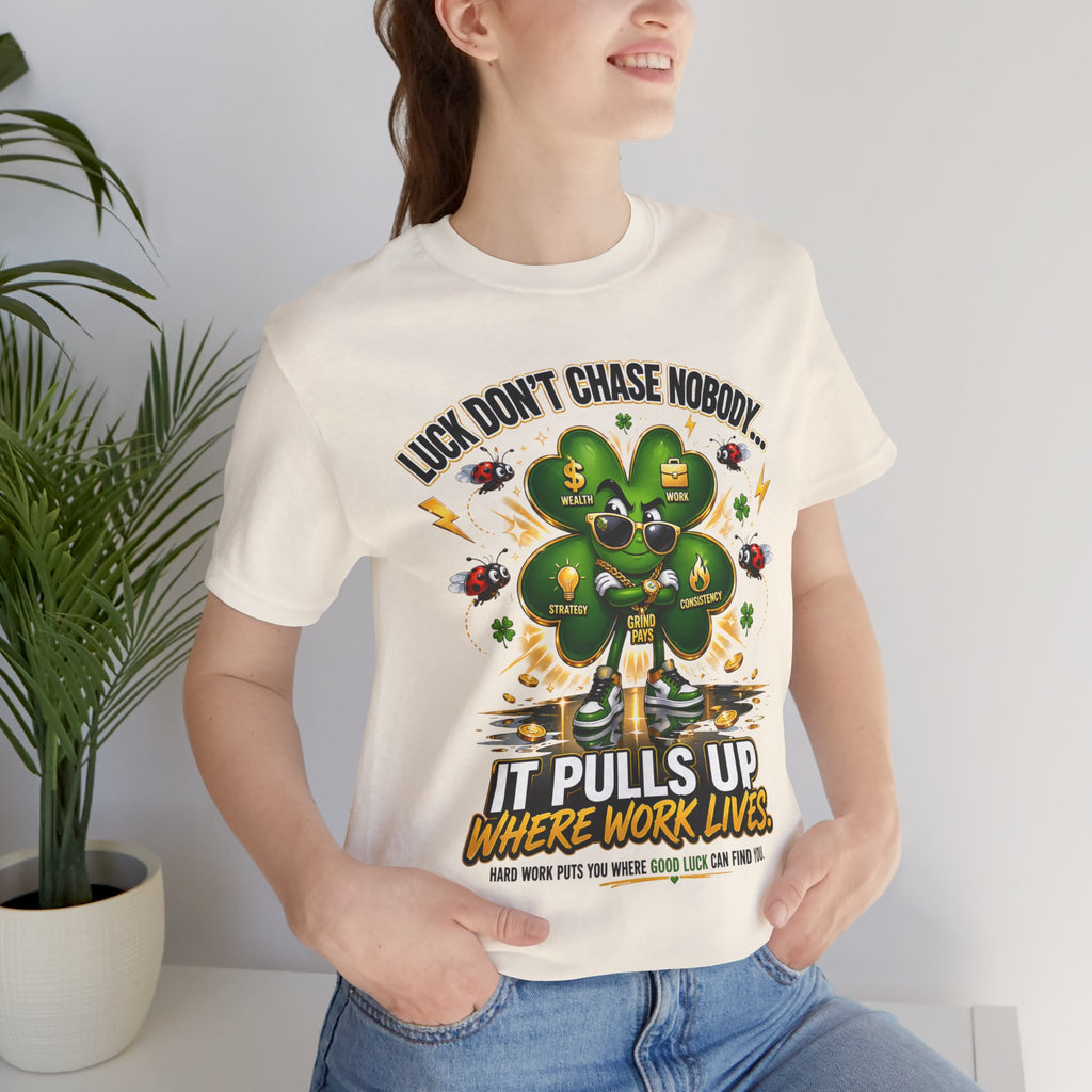 A smiling woman wears the TimeWarp Drip Lucky Clover "Pulls Up Lives" Graphic T-Shirt with clover, coins, and motivational text. She stands indoors near a plant. Available in sizes S-3XL.