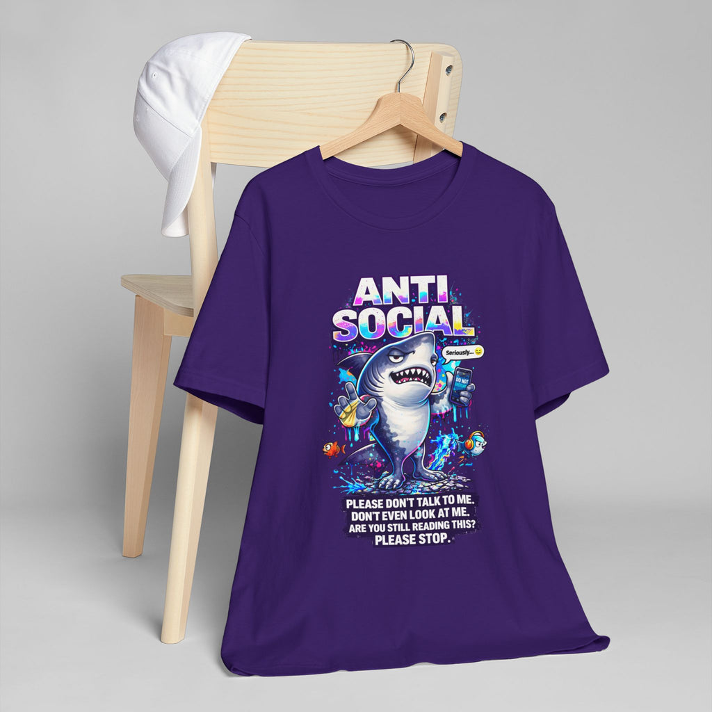 A purple Anti Social Shark T-Shirt — Funny Introvert Graphic Tee with a playful shark design and the text, "Please don’t talk to me. Are you still reading this? Please stop." hangs on a wooden chair. A white cap sits on the backrest.