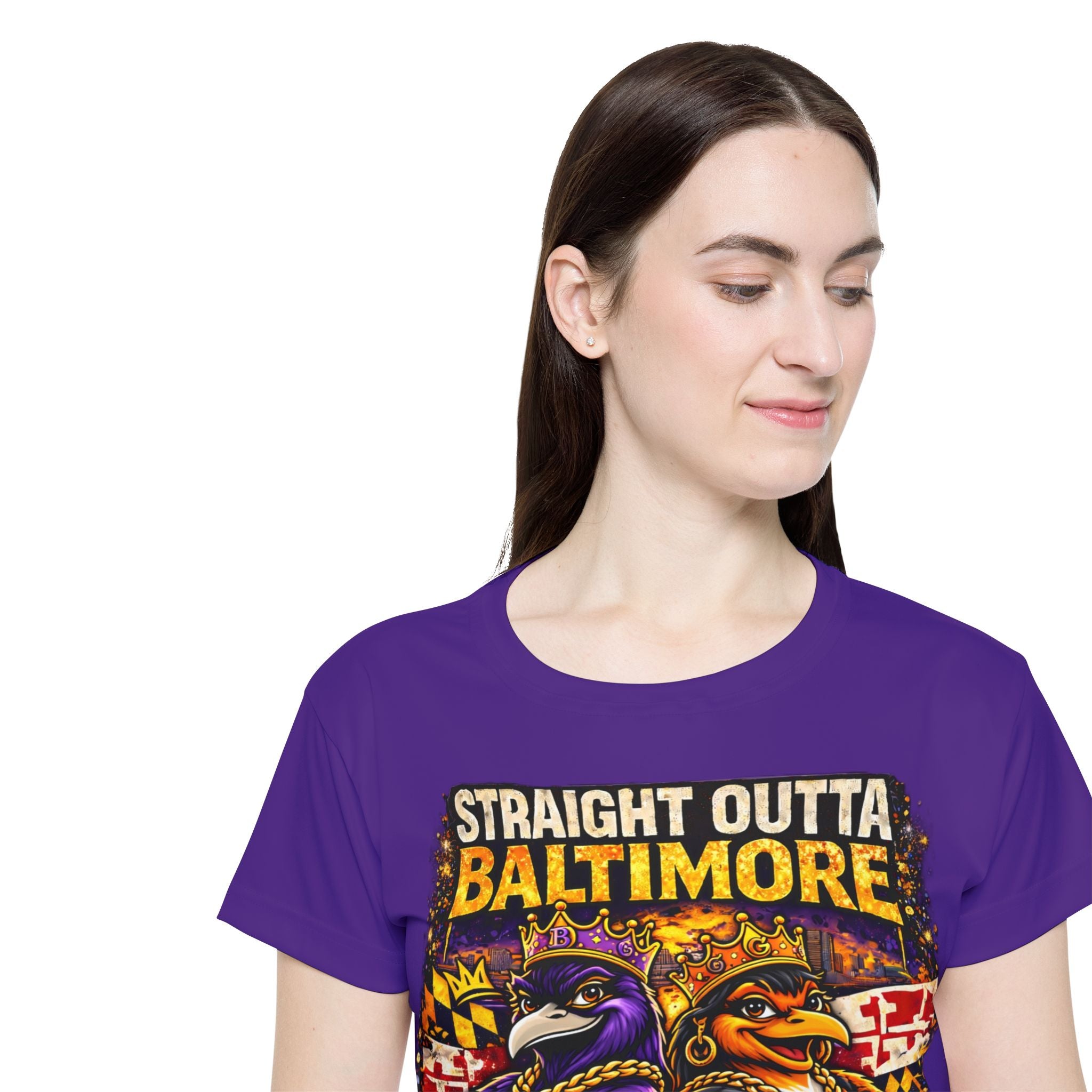 A woman with long brown hair wears the Straight Outta Baltimore — Women’s Sports Jersey, a purple Charm City fan tee featuring two crowned cartoon birds with sports logos. She smiles slightly while looking down against a white background.