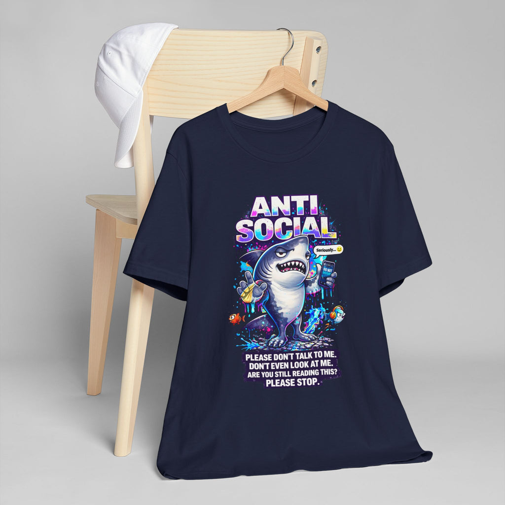 A navy blue Anti Social Shark T-Shirt — Funny Introvert Graphic Tee hangs on a wooden chair with a white cap. It features a cartoon shark holding snacks and the bold text: Please don’t talk to me. Don’t even look at me.