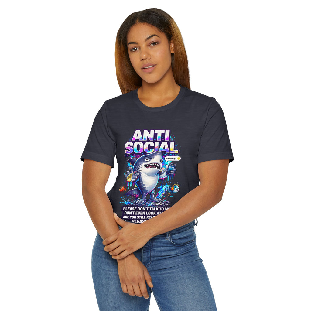 A woman with straight brown hair and blue jeans faces the camera, wearing the Anti Social Shark T-Shirt — Funny Introvert Graphic Tee, a unisex cotton shirt with a cartoon shark and colorful "ANTI SOCIAL" text below.