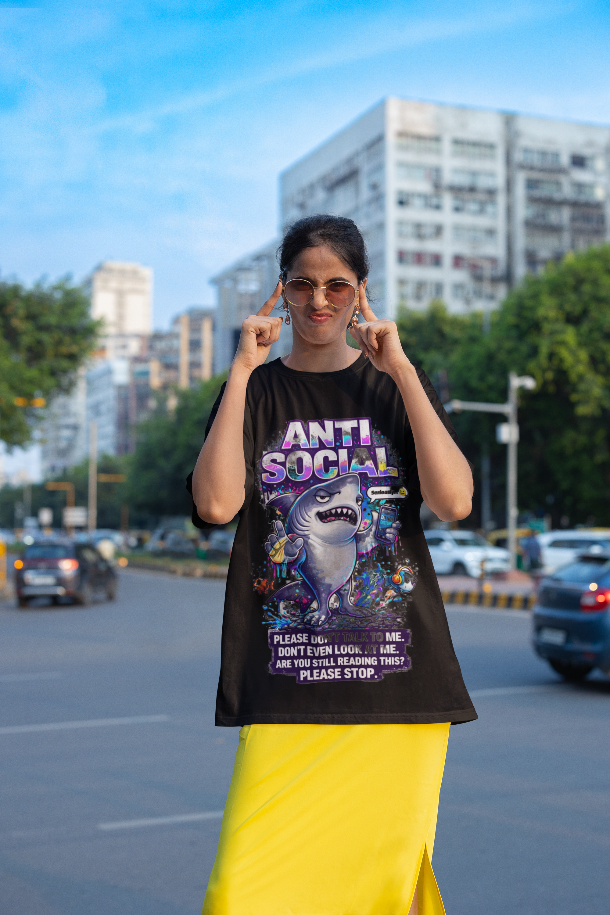 A woman stands on a city street wearing sunglasses, a yellow skirt, and the Anti Social Shark T-Shirt — Funny Introvert Graphic Tee, playfully pulling the sides of her mouth into a smile with her fingers.