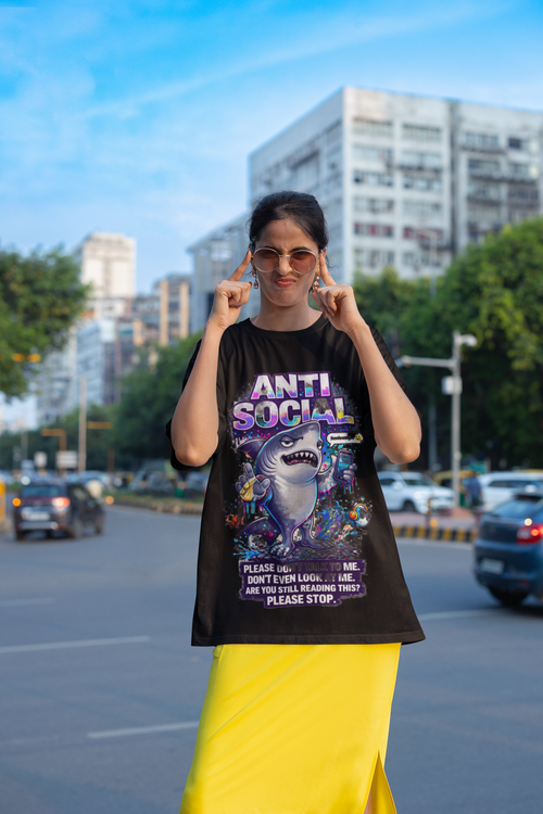 A woman stands on a city street wearing sunglasses, a yellow skirt, and the Anti Social Shark T-Shirt — Funny Introvert Graphic Tee, playfully pulling the sides of her mouth into a smile with her fingers.