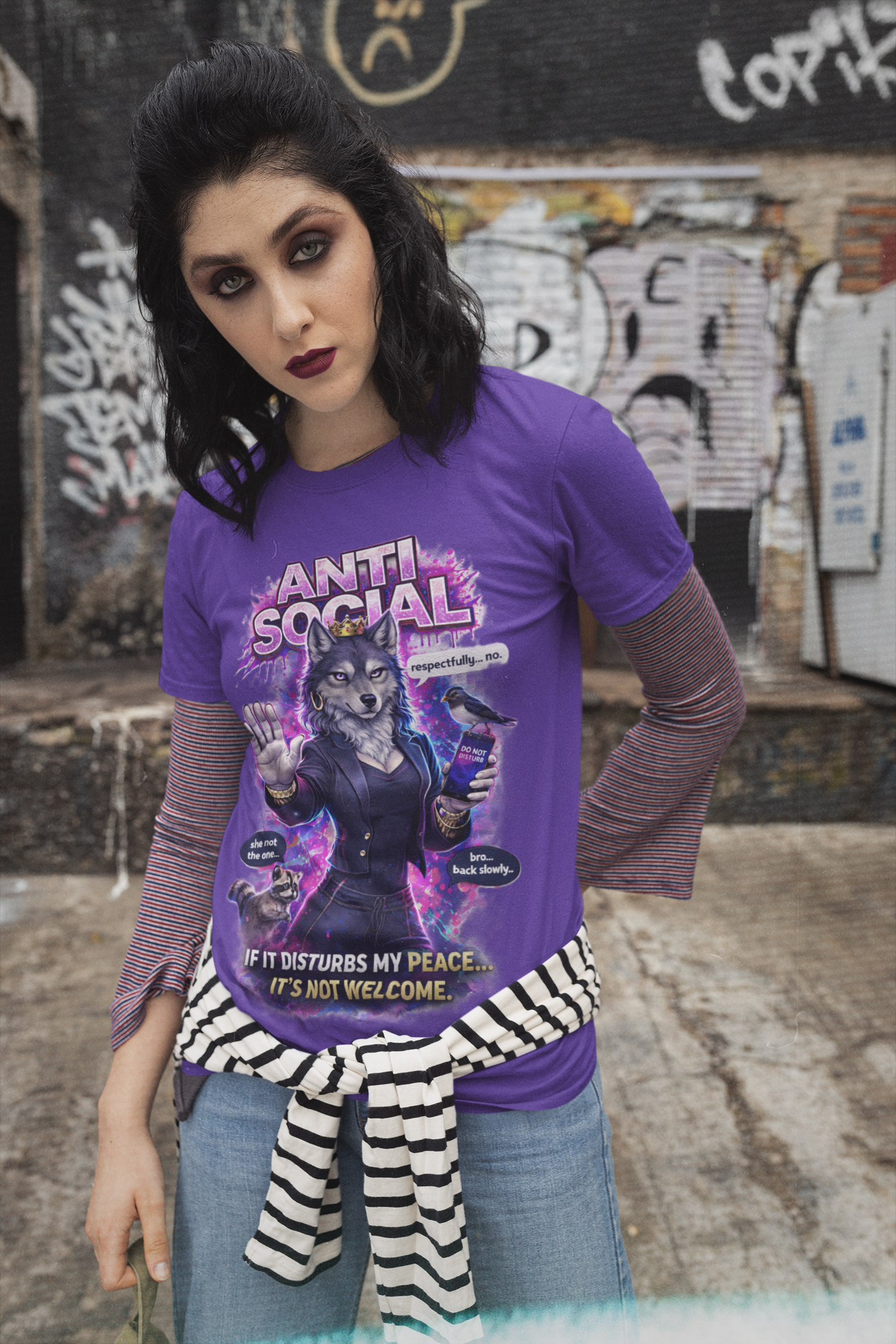 A woman with dark hair and bold makeup stands outdoors by a graffiti wall, wearing an Anti Social She-Wolf T-Shirt—Funny Introvert Graphic Tee over a striped shirt, with a striped sweater tied at her waist.