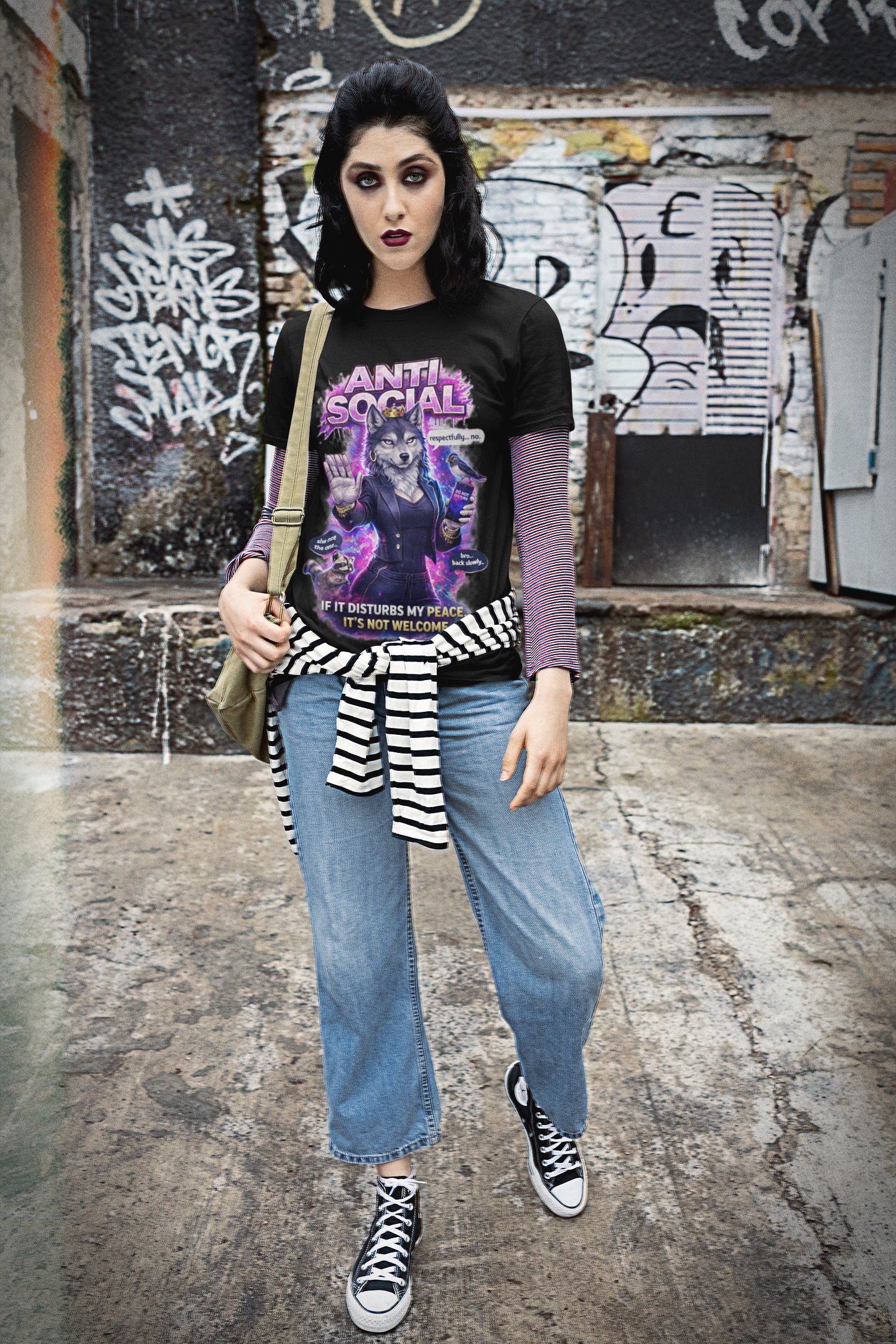 A woman with dark makeup and black hair stands against a graffiti wall, wearing jeans, a striped shirt at her waist, black Converse sneakers, and the Anti Social She-Wolf T-Shirt — Funny Introvert Graphic Tee.