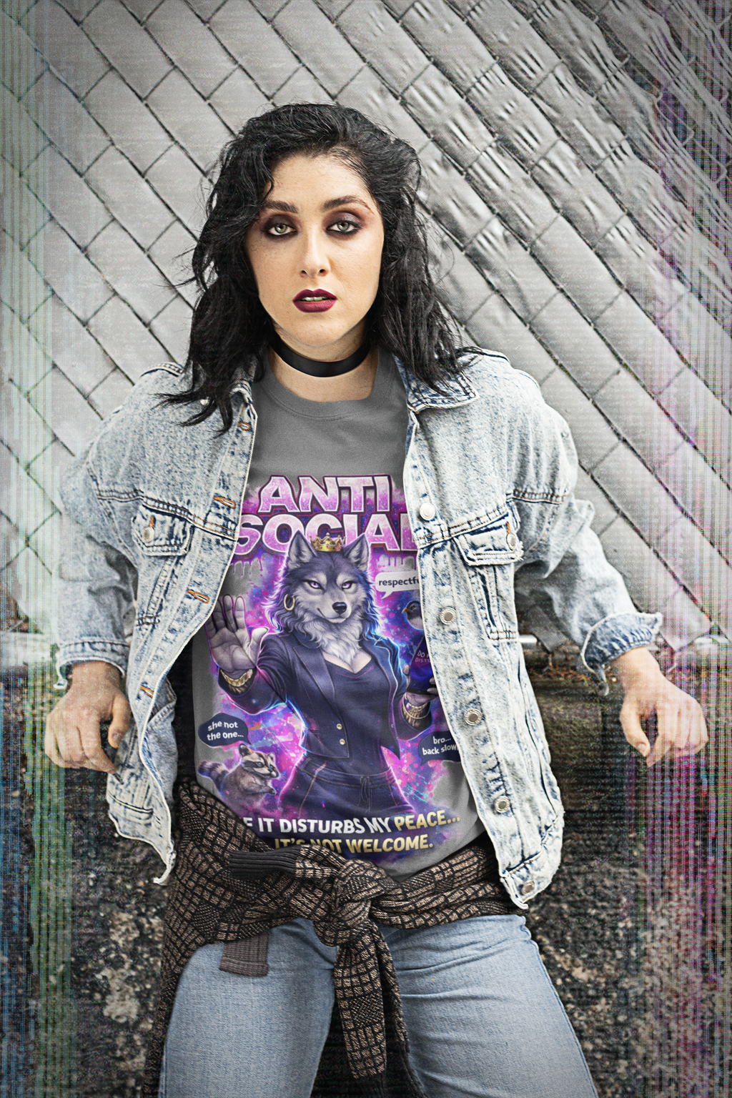 A woman with dark hair and bold makeup stands by a chain-link fence, wearing a jean jacket over the Anti Social She-Wolf T-Shirt — Funny Introvert Graphic Tee. She also sports a plaid shirt tied at her waist and a choker necklace.