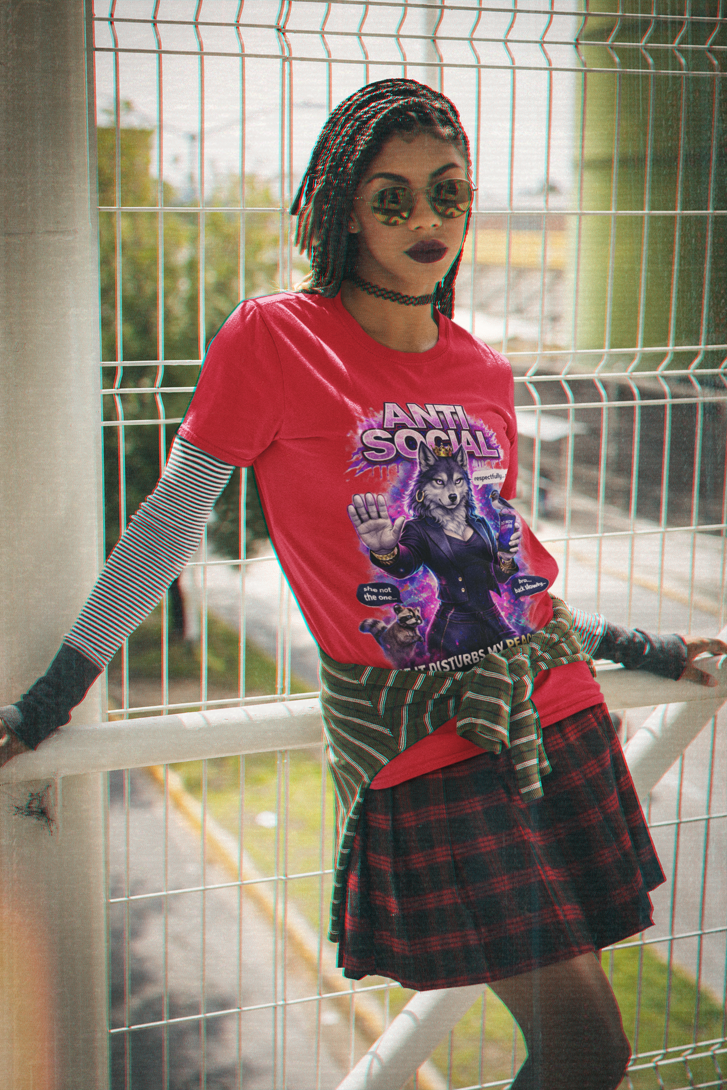 A person with braided hair and bold makeup wears the Anti Social She-Wolf T-Shirt — Funny Introvert Graphic Tee over a striped long-sleeve, paired with a plaid skirt and tie-waist shirt, standing by a metal fence outdoors.