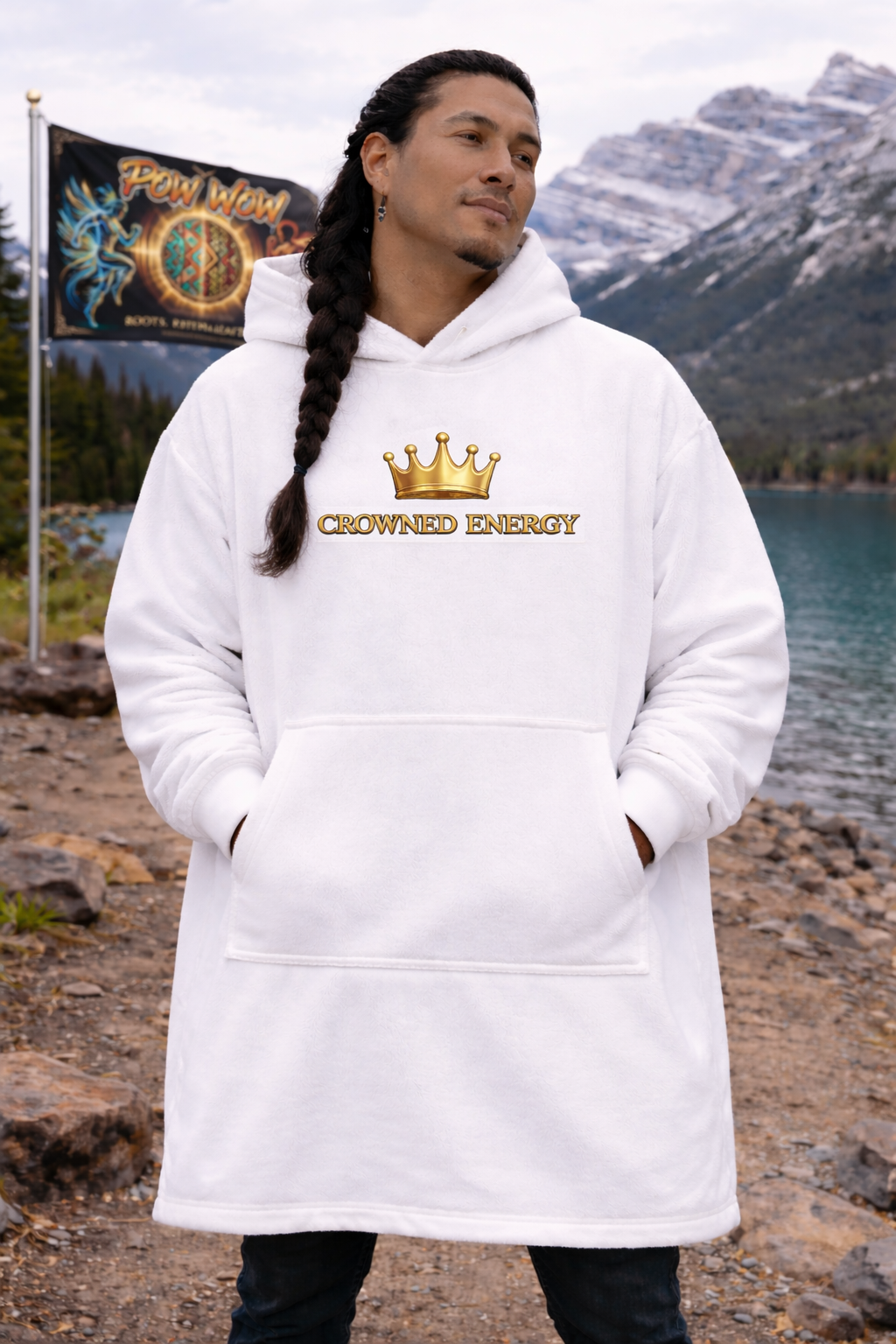 A person with long braided hair stands outdoors by a lake and mountains, wearing the "Crowned in Every Era" Royal Chess Graphic (White) Hooded Blanket. A Pow Wow banner is visible in the background. Sleeve length is standard.