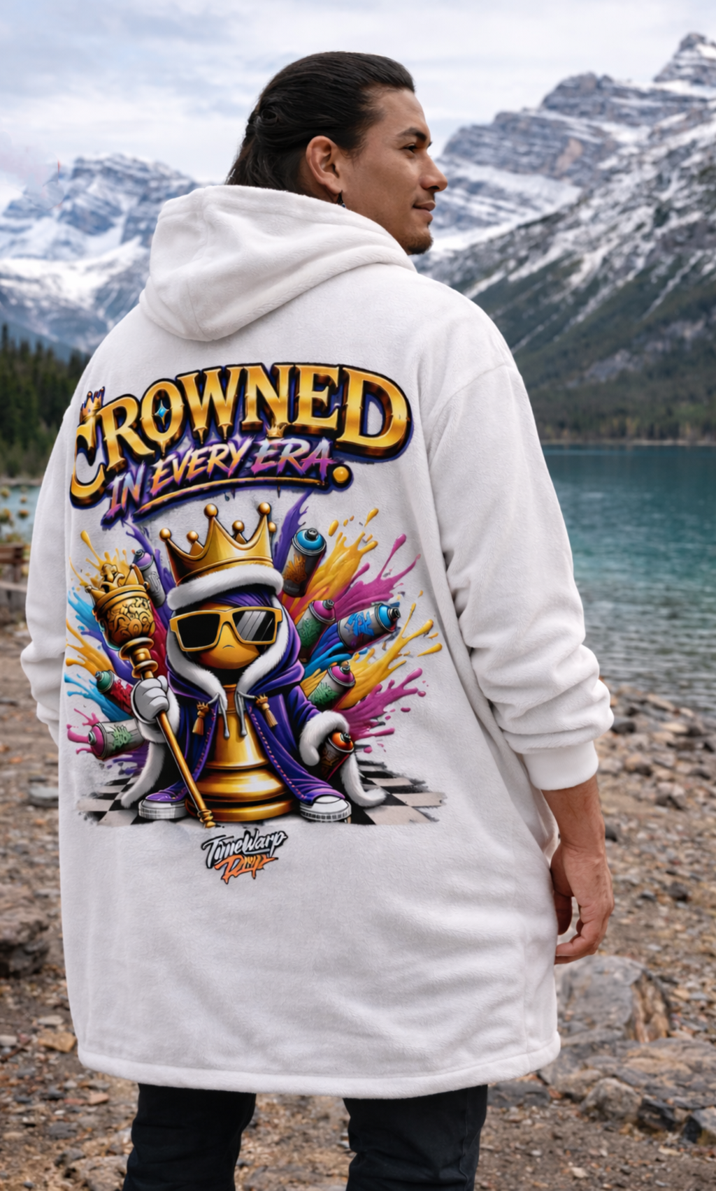 A man stands by a mountain lake in the "Crowned in Every Era" Royal Chess Graphic (White) Hooded Blanket, featuring a colorful cartoon king, paint cans, and text on the back. Snowy mountains and trees are behind him.