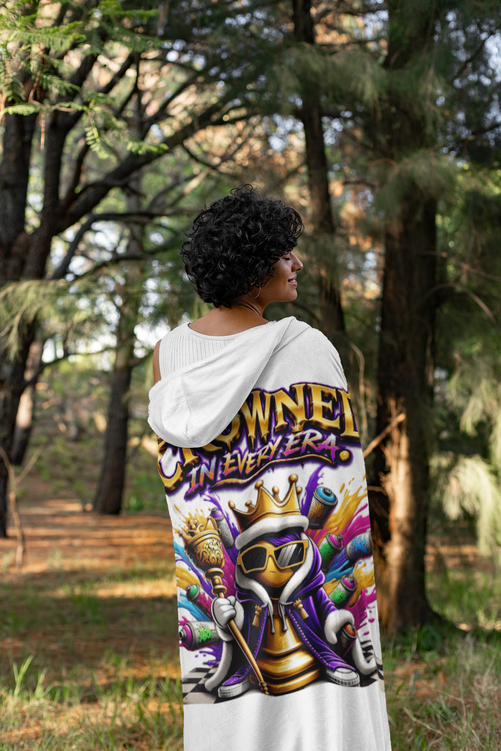A person with short curly hair stands outdoors wearing the "Crowned in Every Era" Royal Chess Graphic (White) Hooded Blanket, featuring a colorful crowned skull with sunglasses, spray paint cans, and bold text.