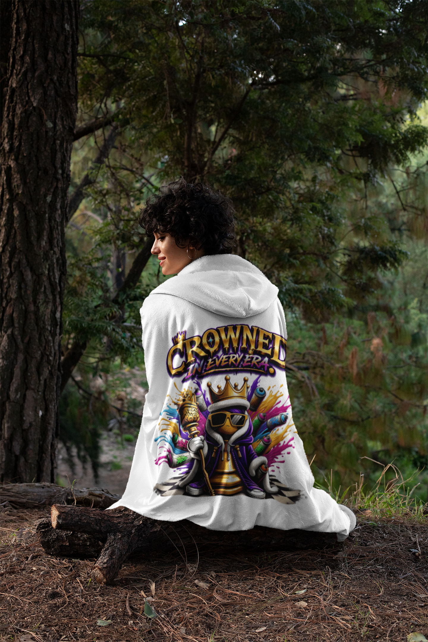 Outdoors on a log, a person with short curly hair wears the "Crowned in Every Era" Royal Chess Graphic (White) Hooded Blanket, featuring a crowned skeleton and bold lettering on the back, with forest trees behind them.
