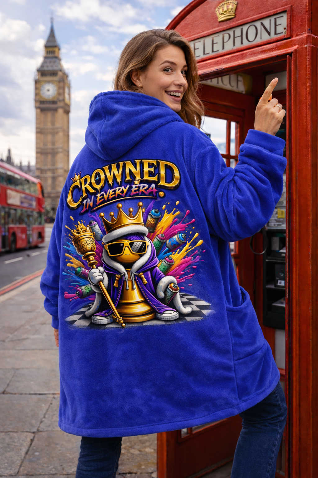 A woman smiles in London near a red phone booth, Big Ben, and buses, wearing the "Crowned in Every Era" Royal Chess Graphic Hooded Blanket in royal blue, featuring a colorful cartoon king and the phrase on the back.