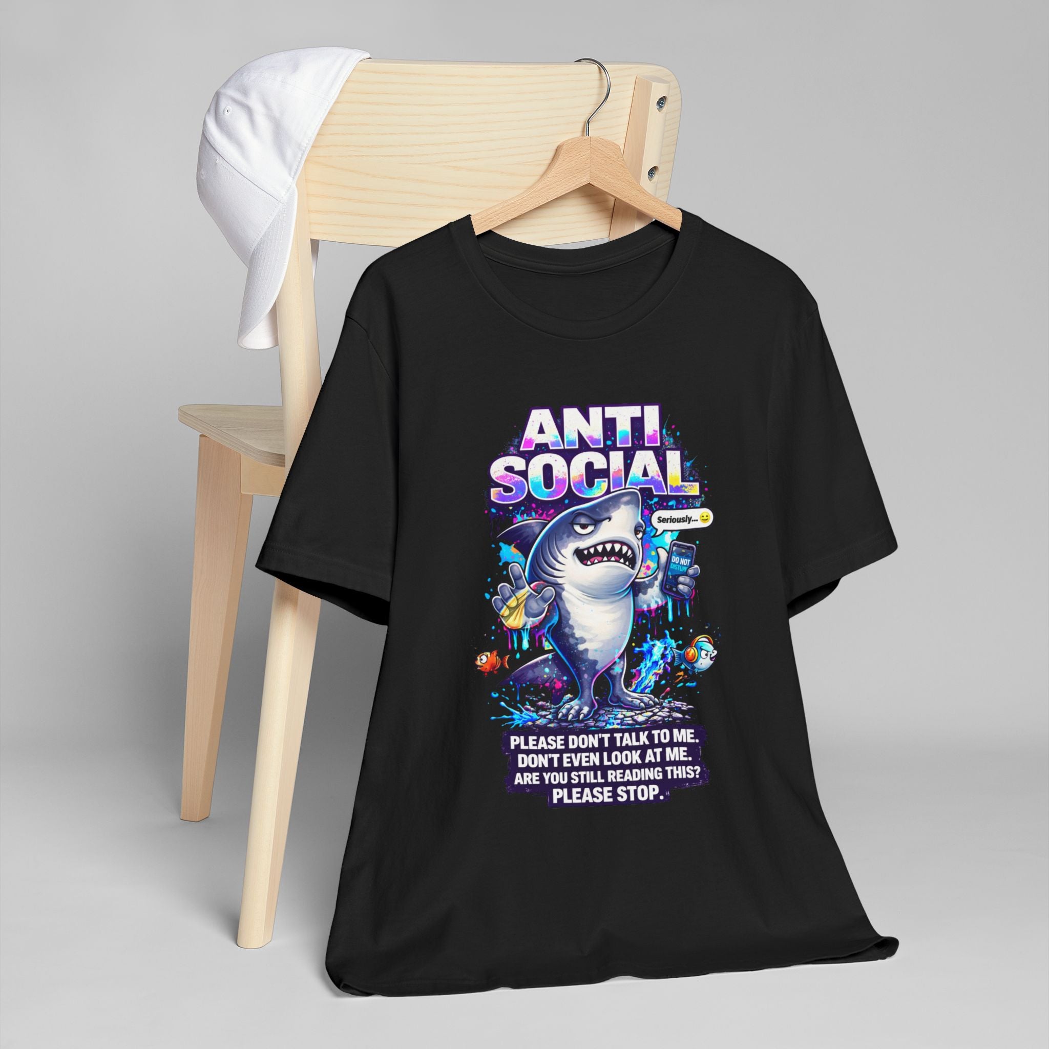 A black unisex cotton Anti Social Shark T-Shirt — Funny Introvert Graphic Tee, featuring a cartoon shark and playful phrases, hangs on a wooden chair with a white cap resting nearby.