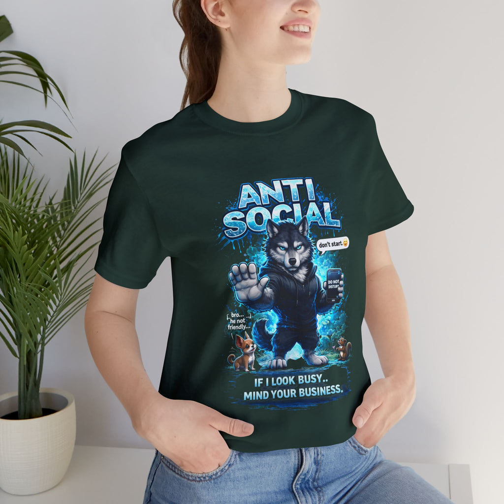 A smiling person wears the Anti Social Wolf T-Shirt — Funny Introvert Graphic Tee a dark green streetwear tee featuring a cartoon wolf with the text "If I look busy, mind your business," two animals, and stands by a potted plant.