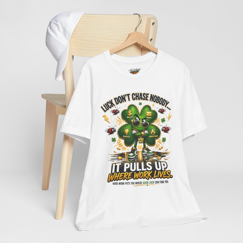 The TimeWarp Drip Lucky Clover "Pulls Up Lives" Graphic T-Shirt, available in sizes S–3XL, sports a bold green four-leaf clover and motivational quote. It’s displayed draped over a wooden chair with a white cap hanging from the side.