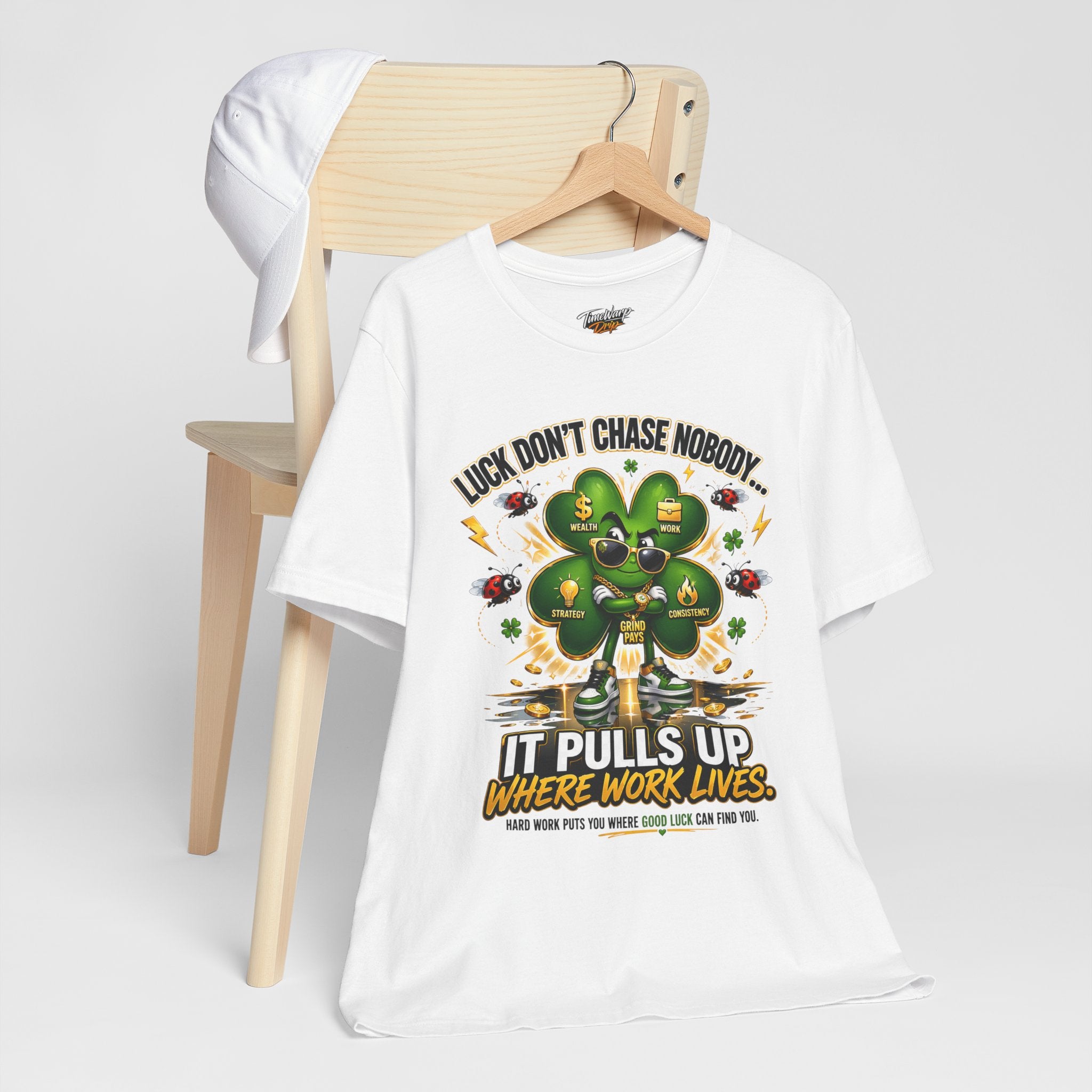 The TimeWarp Drip Lucky Clover "Pulls Up Lives" Graphic T-Shirt, available in sizes S–3XL, sports a bold green four-leaf clover and motivational quote. It’s displayed draped over a wooden chair with a white cap hanging from the side.