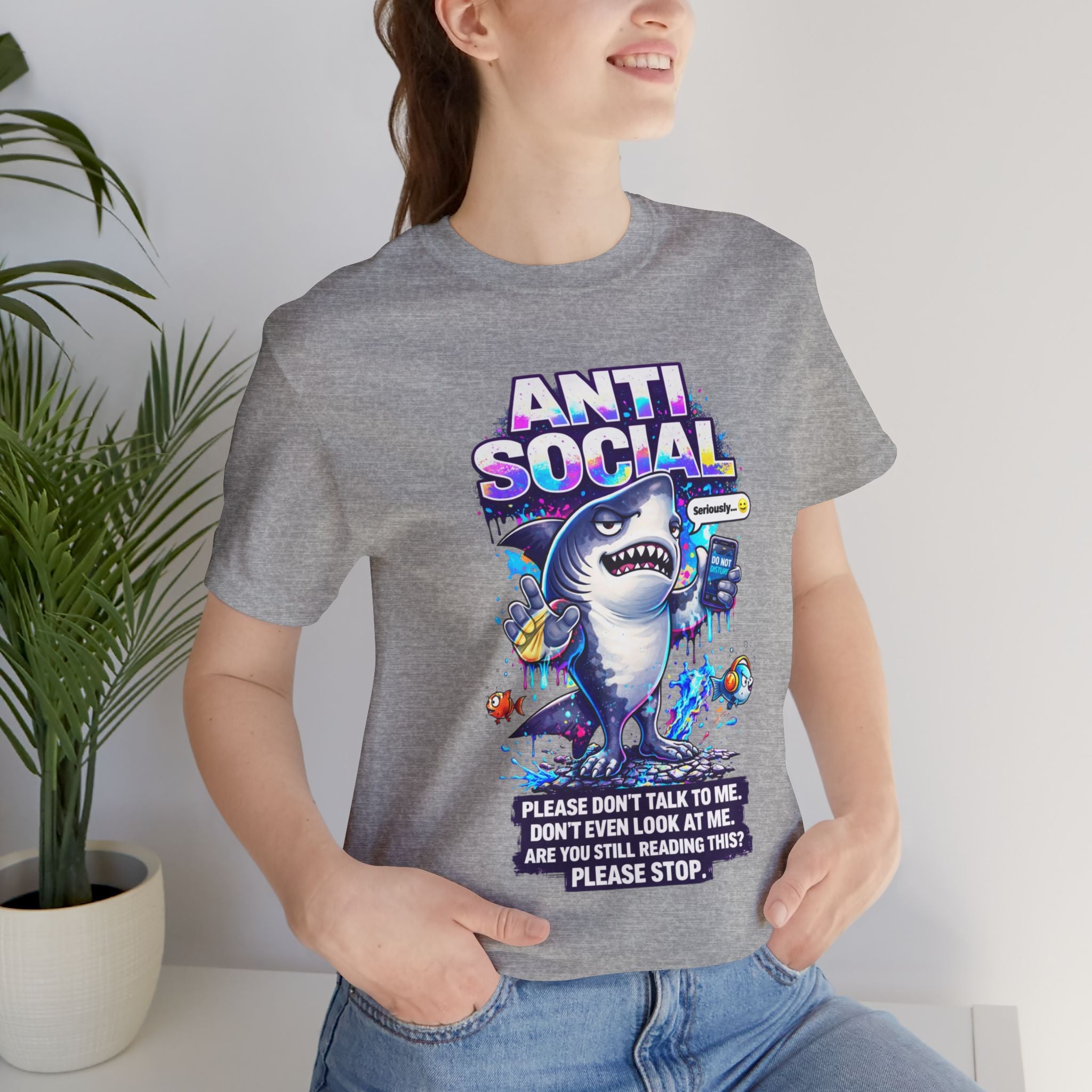 A smiling person wears the Anti Social Shark T-Shirt — Funny Introvert Graphic Tee, showing off its playful shark graphic and bold lettering indoors, with a leafy plant adding to the cozy vibe.